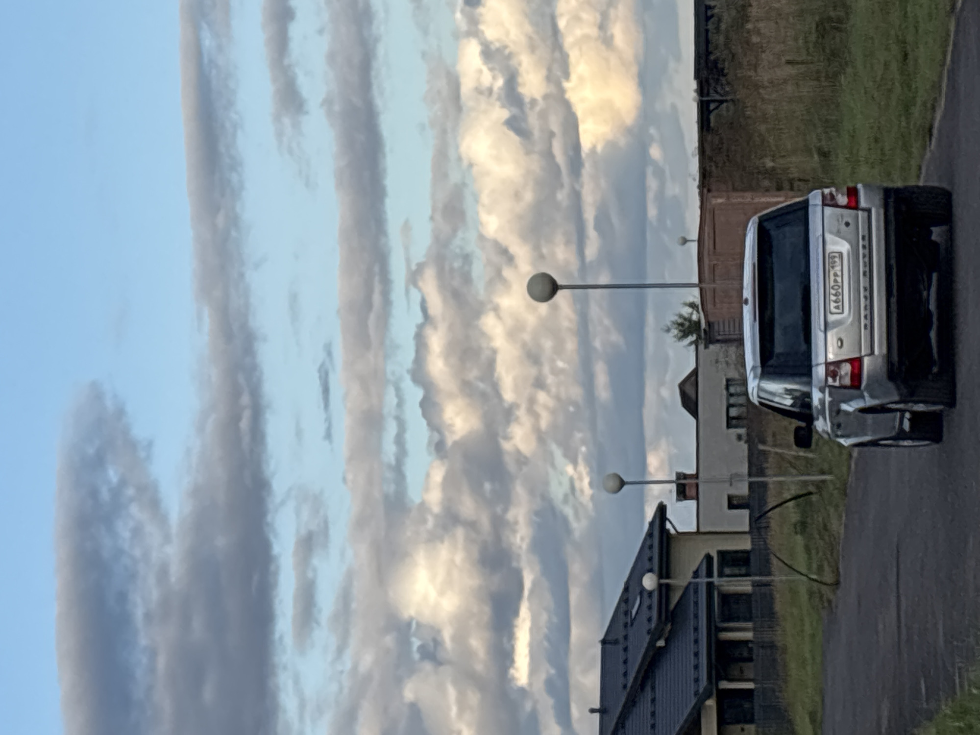 A silver Range Rover is parked on a paved road in front of modern residential buildings and a lot of dry vegetation. The sky above is filled with dramatic clouds. The license plate of the Range Rover is visible and reads "A660PP 199". The scene appears to be in Zvenigorod, Russia.