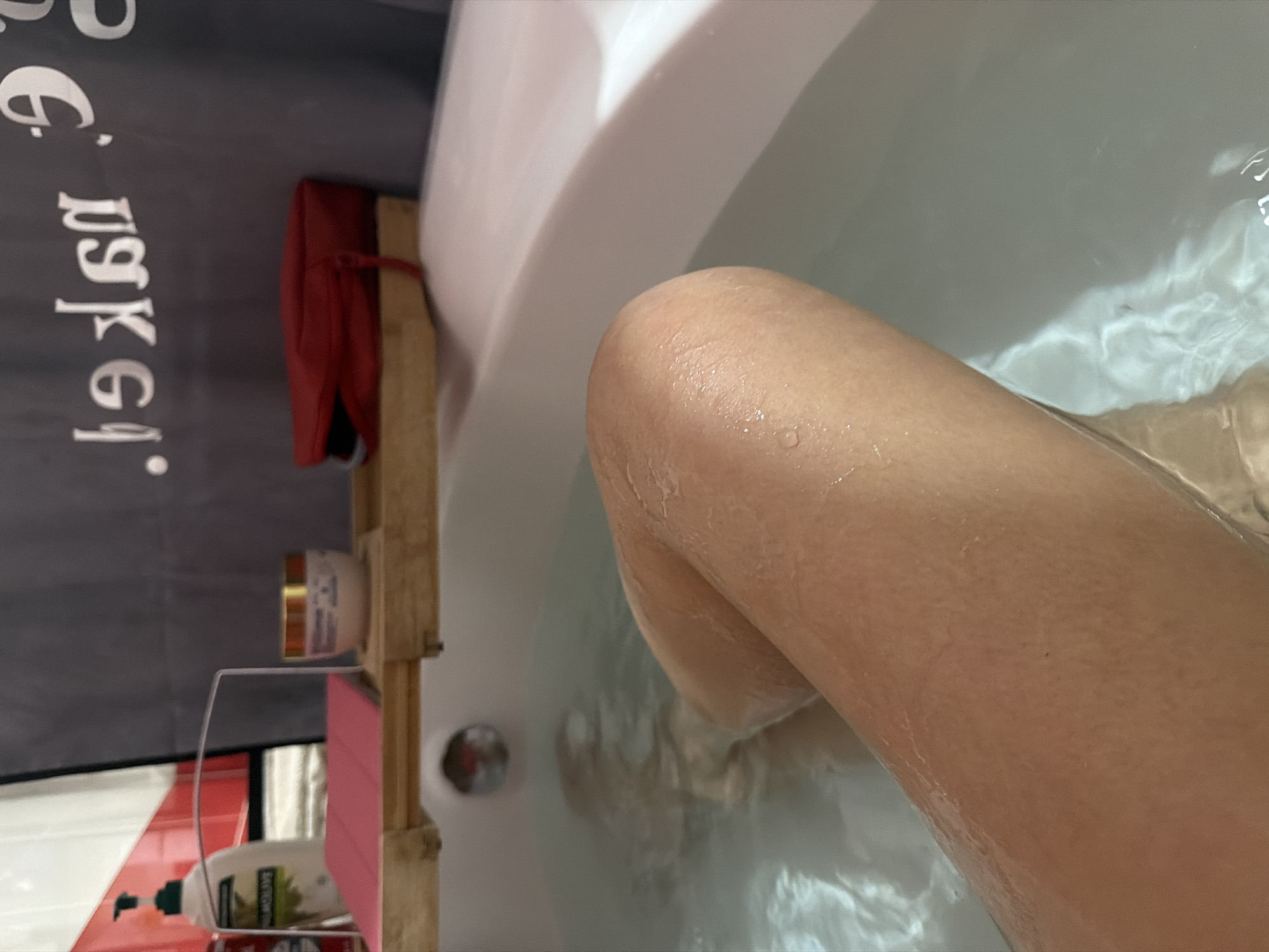 The image shows a partial view of a person in a bathtub filled with water. The individual's leg is prominently visible, with water droplets on the skin. A wooden bathtub caddy holds various items, including a red bag, a jar, and a bottle of soap. The caddy is positioned next to a gray shower curtain with white lettering. The location is identified as Большая Шильна, Russia.