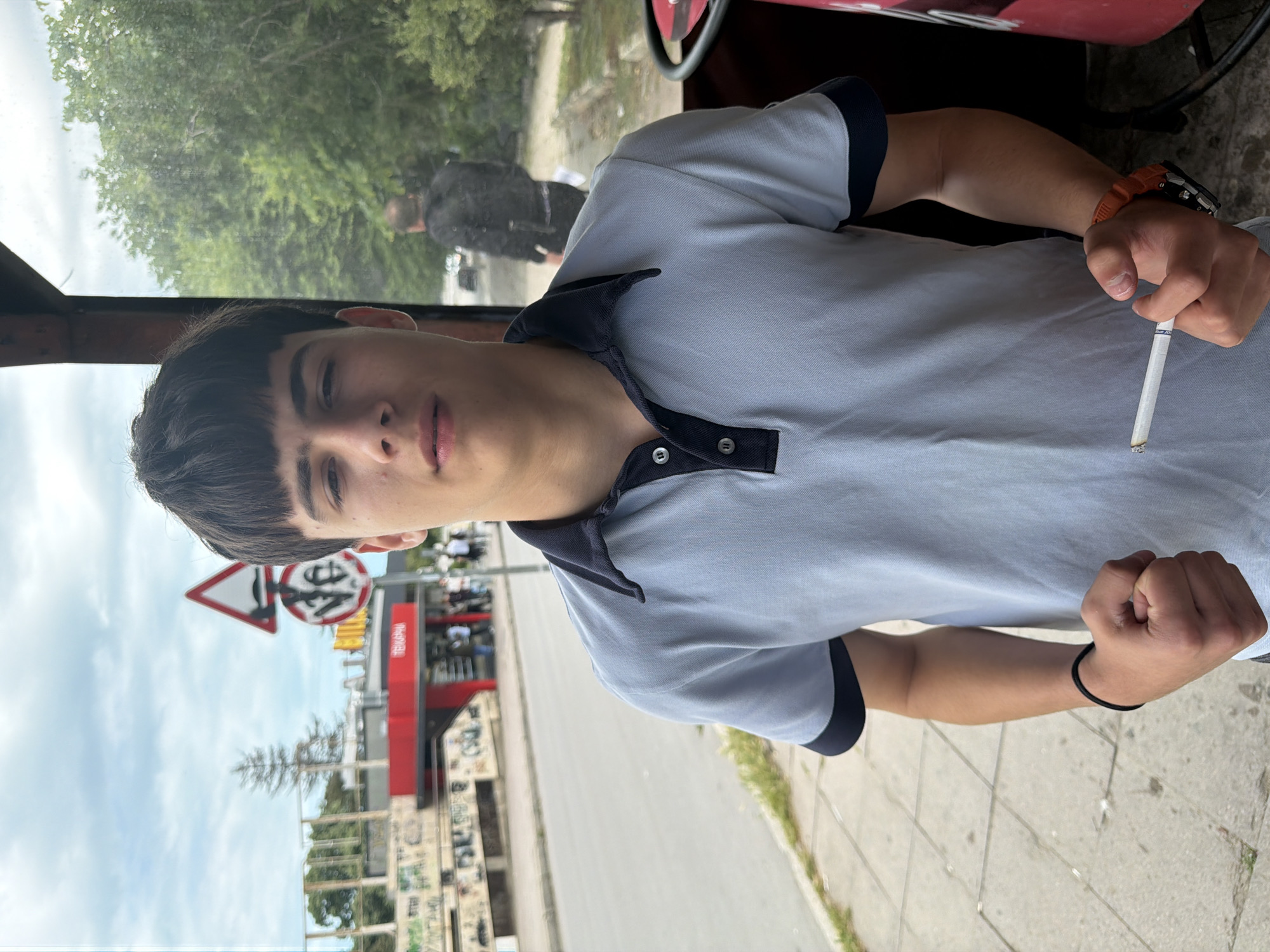 A young man stands, holding a lit cigarette in his right hand, wearing a gray polo shirt with a dark blue collar and sleeve trim. He has short dark hair and is wearing a watch on his right wrist and a bracelet on his left wrist. He appears to be inside some form of shelter or vehicle, as suggested by the roof and window. The location is Varna, Bulgaria, evident from the "ТЕХНИКА" sign and distinctive road signs visible in the background. The scene includes an urban street setting with buildings, street signs, and a visible road.