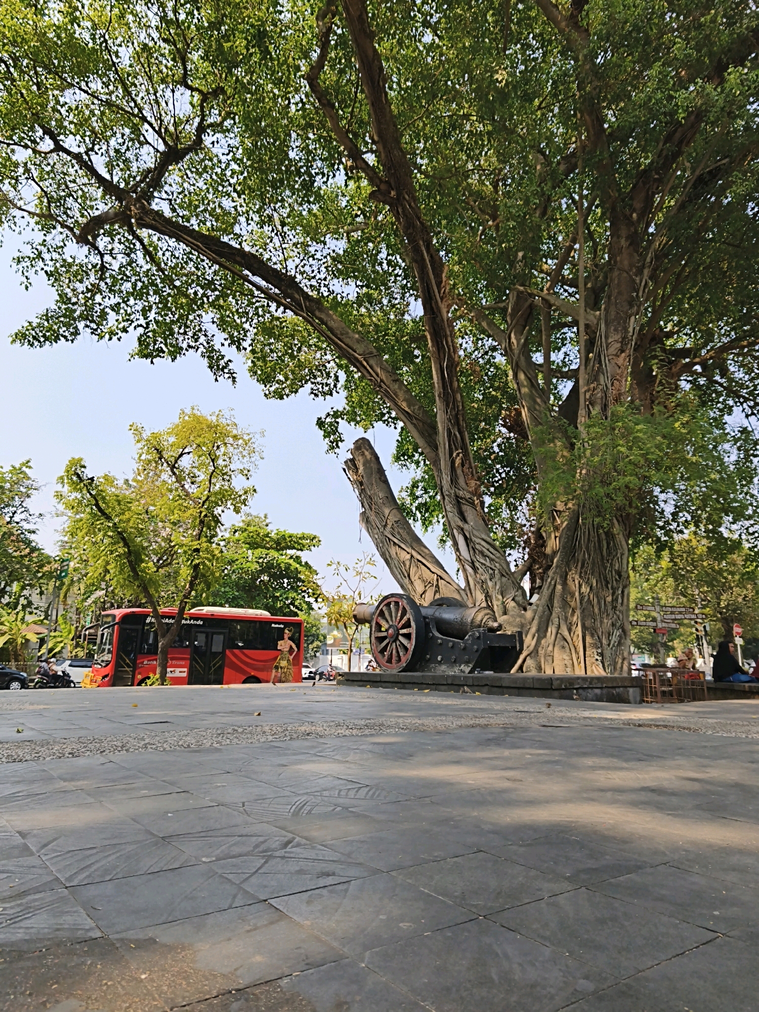 This image captures an outdoor scene in Surakarta, Indonesia, likely within or near a public park or square, characterized by a large, old banyan tree with extensive aerial roots. In the foreground, a stone-tiled plaza stretches out, with some areas showing wear and tear and scattered debris. To the right of the plaza, a vintage cannon is prominently displayed, resting on a stone base. Behind the cannon and partially obscured by the banyan tree's trunk, a woman in a dark headscarf and blue clothing is seated on a bench, while another person in white is visible nearby.
To the left of the banyan tree, a red bus with "Batik Trans" branding is parked. The bus features an advertisement of a woman in a traditional patterned dress on its side. Several vehicles, including cars and motorcycles, are visible on the street behind the bus. A few individuals can be seen near the vehicles, with one person standing in front of the bus.
The background is filled with lush green trees, suggesting a verdant environment within the city. The sky is clear and bright blue, indicating a sunny day. The overall atmosphere appears to be that of a historical or cultural site within Surakarta, with the presence of the cannon and the ancient tree contributing to a sense of heritage. The lighting suggests it is late afternoon, with long shadows cast across the plaza.