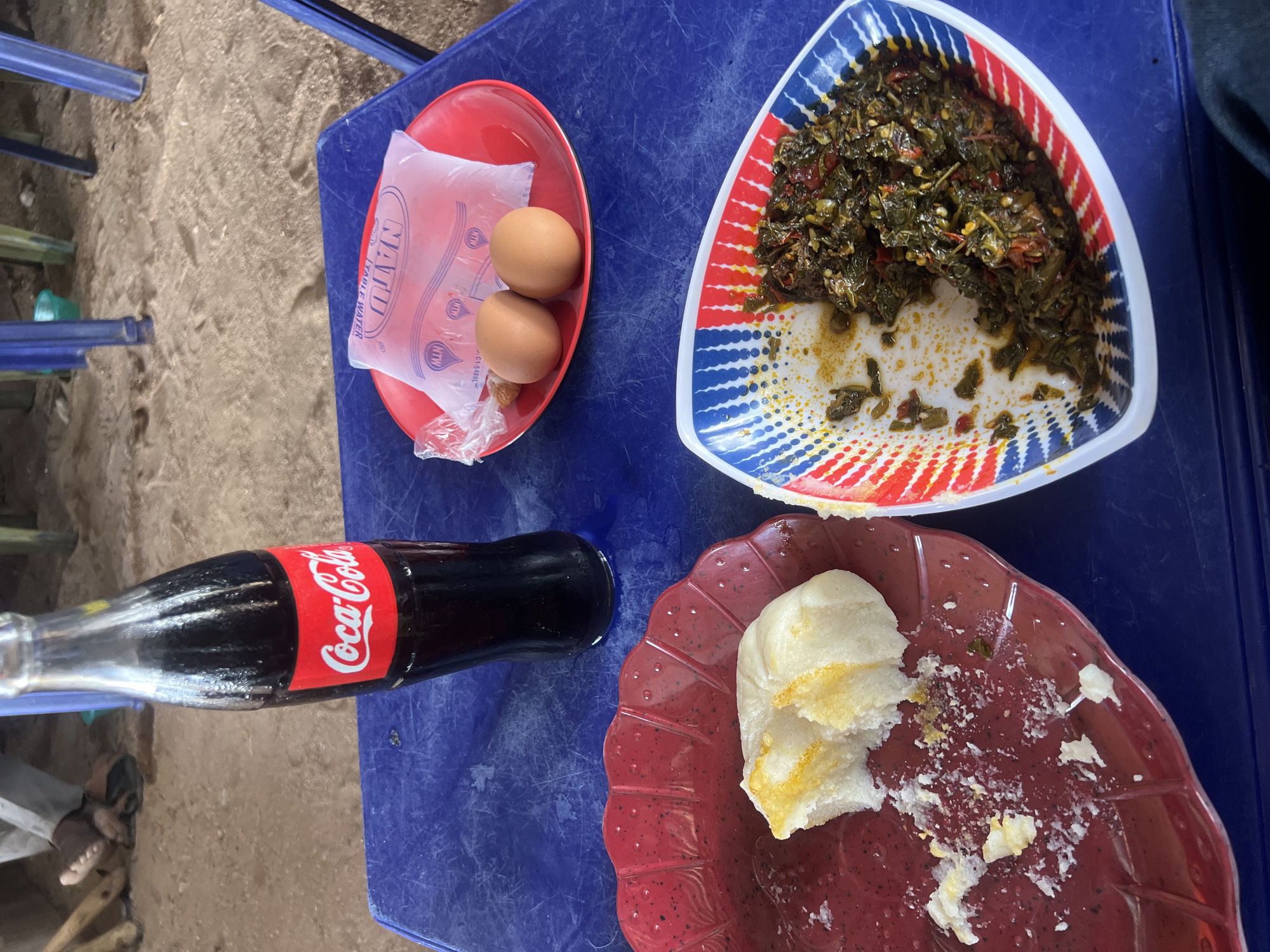 The scene captures a meal presented on a scratched blue table, situated in an outdoor or semi-outdoor dining area, consistent with local eateries in Jalingo, Nigeria. A dark blue fabric, indicative of a person's leg or lap, is visible at the bottom right. The table hosts a red, scalloped plate with a partially consumed white starchy food, possibly *fufu*, displaying yellow sauce remnants and crumbs. A standard glass bottle of Coca-Cola with its red branding label stands upright. To its right, a plain red plate holds two raw brown eggs and a clear plastic sachet labeled 'NTW TABLE WATER', featuring small 'MTN' logos and a batch identifier 'CT.34BBL'. Further right, a white bowl with red and blue stripes contains a partially eaten dark green leafy stew with red pepper fragments. The background consists of sandy ground with several blue and green metal furniture legs. A pair of bare feet, wearing brown sandals, are discernible on the sand in the far left background.