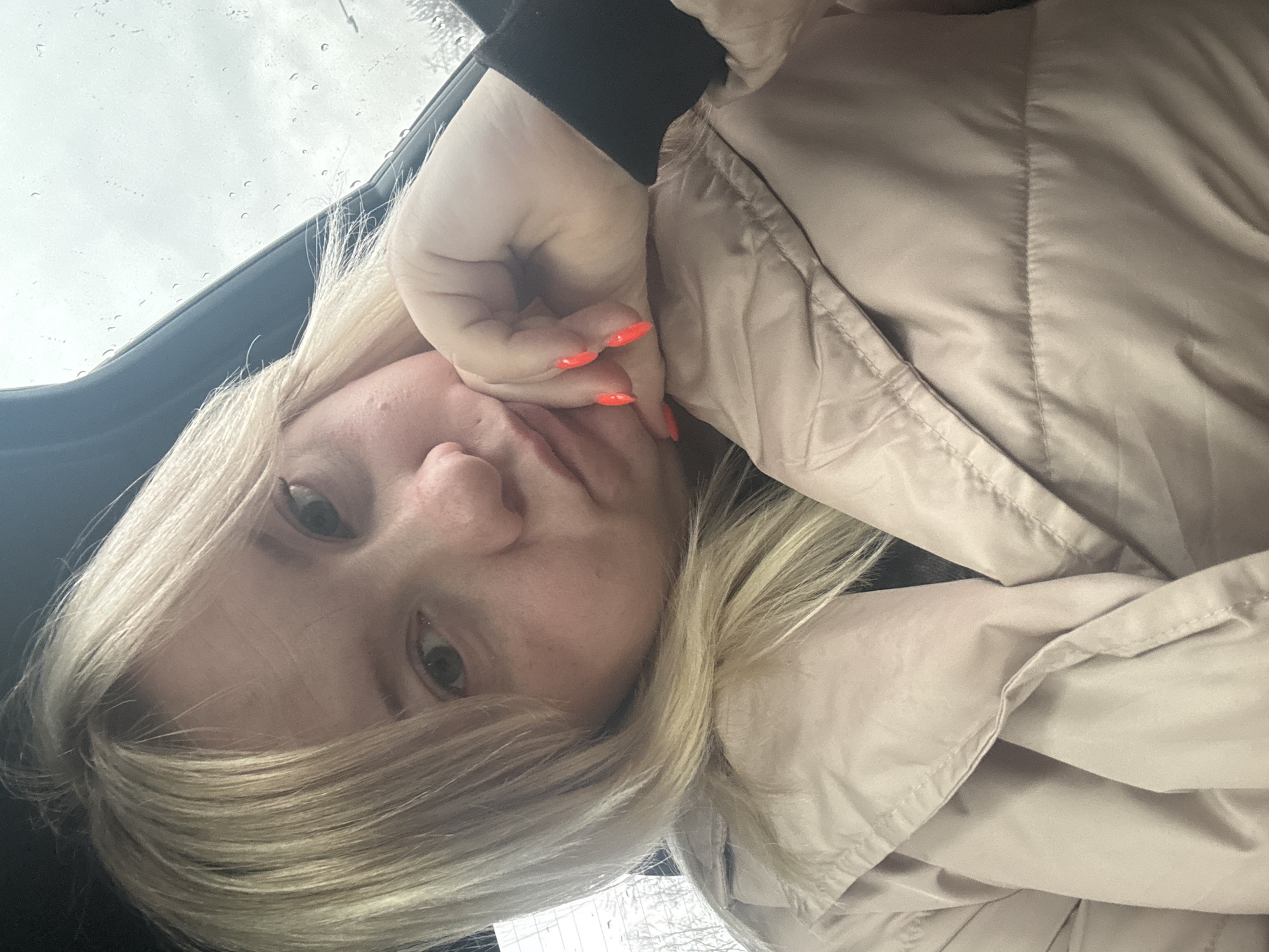 A close-up, slightly rotated portrait captures a young woman inside what appears to be a vehicle. She has blonde hair and a fair complexion, looking directly into the camera with a neutral, perhaps pensive, expression and a slight pout. Her right hand is resting against her cheek and chin, partially obscuring her mouth. Her fingernails are painted a vibrant orange or coral color. She is wearing a light beige or tan puffy jacket, visible on her upper body, and a black sleeve is seen beneath her hand.
Through the window behind her, an overcast sky is visible, marked by numerous raindrops, indicating rainy or recently rainy weather. The dark frame of the car window is noticeable in the upper left. The outdoor background beyond the window is indistinct and blurry. The scene suggests a casual moment, possibly a selfie, taken during the daytime in a car, with the weather being a significant detail. No text is visible.