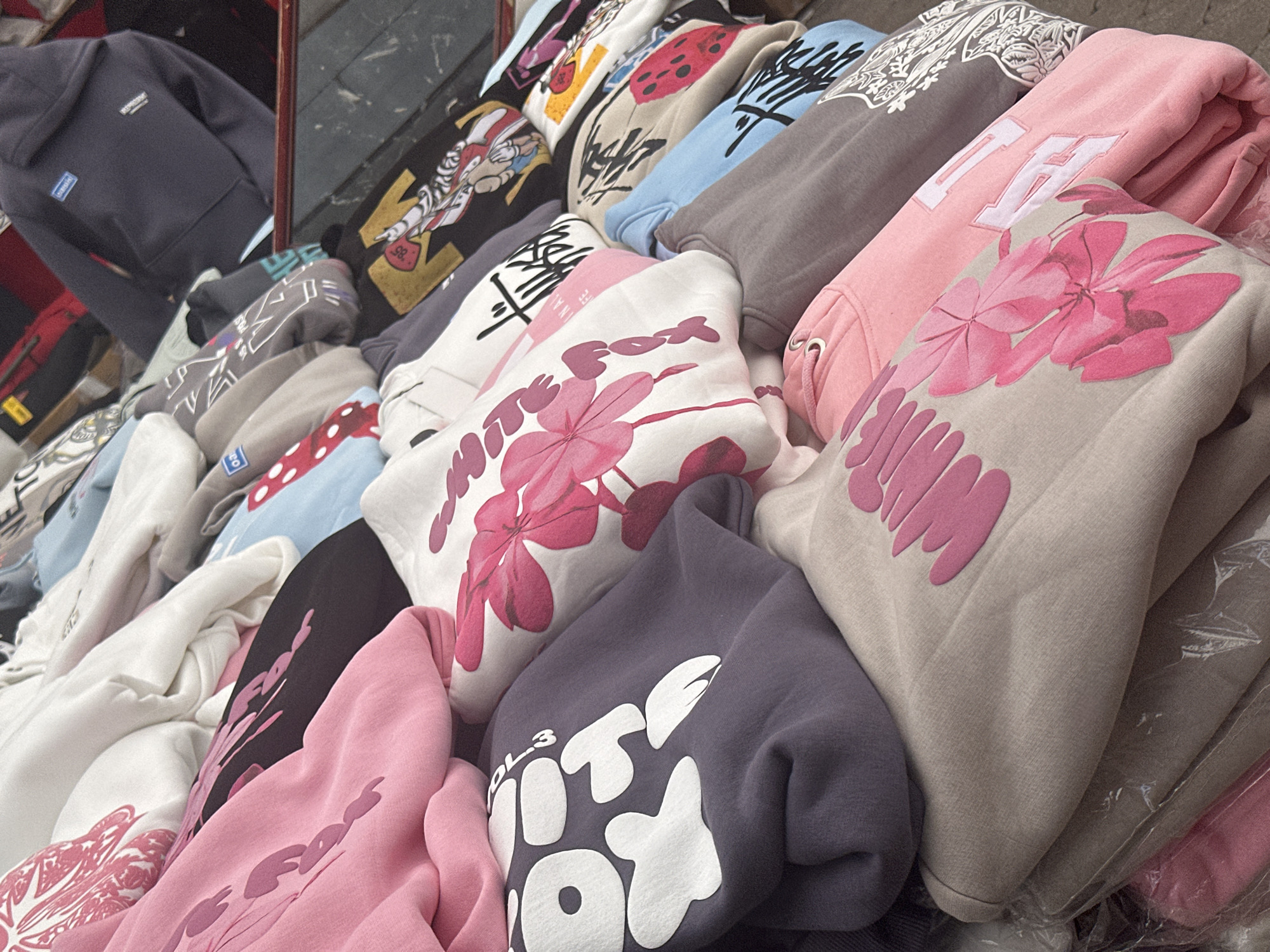The image shows a pile of folded clothes, mostly hoodies with various designs and colors, stacked seemingly on a vendor's display. The clothing includes designs featuring floral patterns, lettering, and character art. The stack includes shades of gray, pink, blue, white, and black. The setting appears to be a market or a retail location. Based on the prompt, this image would have been taken in the city of Fethiye, Turkey. There is no evidence of people or action within the scope of the image.