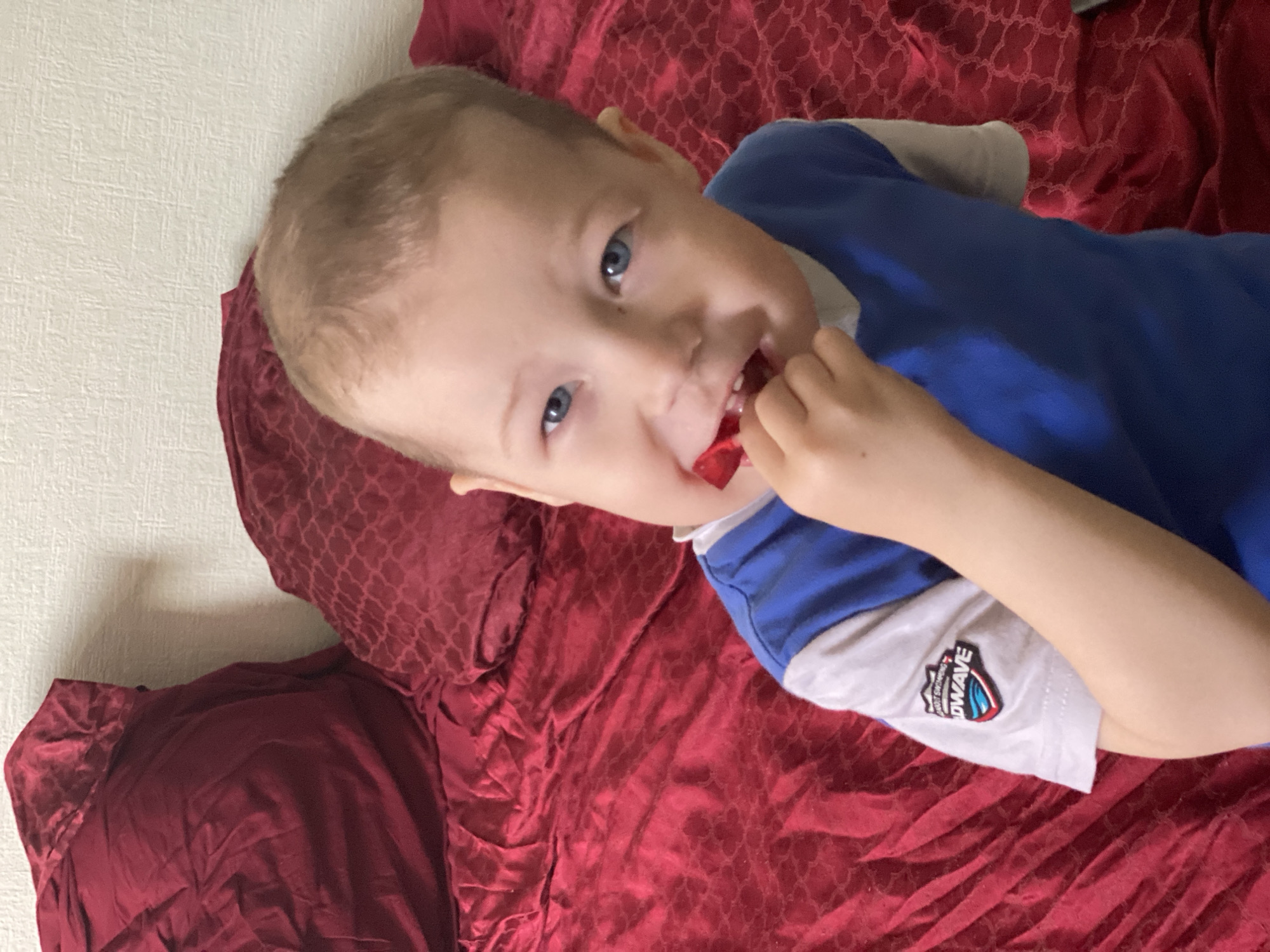 A young child with blue eyes is lying on a red bedspread and holding a red object in his mouth. The child is wearing a blue and white shirt. The background includes a wall and the patterned red bedspread. The location is not discernible from the image and therefore cannot be specified as Sochi, Russia.