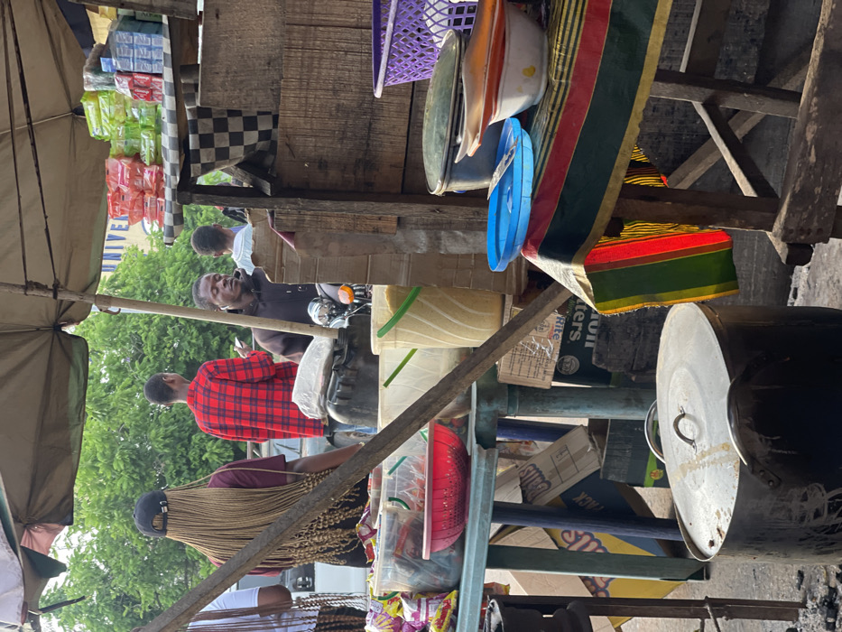 This image captures a vibrant and busy outdoor market scene, likely taken during the daytime. The perspective is from within or very close to a market stall, looking outwards towards people and other stalls. The scene is characterized by a mix of makeshift wooden structures, goods for sale, and several individuals.
In the foreground on the left, a large, light-colored umbrella or canopy provides shade. Below it, in the lower left, a person is visible from the back, wearing a black cap and sporting long, light-colored braids. Next to them, further into the mid-ground, another person is seen from the back, wearing a red and black plaid shirt.
Central to the mid-ground, a man with dark skin is seated on what appears to be a motorbike, or at least has a motorbike in front of him. He is wearing a dark-colored shirt and has an animated expression, with his mouth open, looking towards the right side of the frame. Behind him, another person in a lighter-colored shirt is partially visible. In the background, beyond the people, there are green trees and a building with the word "UNIVERSITY" partially visible on its facade.
The right side of the image is dominated by a market stall setup. It consists of rough wooden shelves and supports packed with various items. Towards the top right, a purple plastic basket, along with white, orange, and blue plastic bowls or basins, are hanging or displayed. Below these, a colorful fabric with distinct red, green, yellow, and black stripes is draped over a wooden beam.
On the shelves and surfaces of the stall, numerous items are stacked and displayed. These include brightly colored packets of goods, possibly snacks or small household items, in green, pink, and blue clear plastic packaging. A black and white checkered cloth is also visible on one of the wooden surfaces. Lower down in the stall area, there are yellow and clear plastic buckets, one with a green handle, and a red plastic colander or basket. Large metal pots with lids are prominently placed in the lower right corner, suggesting cooking or serving food. Several cardboard boxes are stacked and scattered around the stall, with visible text including "BUTTERS," "Galls," "PACK," and smaller text that seems to include "Rite."
The overall impression is one of a lively, functional marketplace with goods readily available and people moving through or engaging with the vendors. The lighting suggests a bright day, possibly with some cloud cover or shade from the canopies.