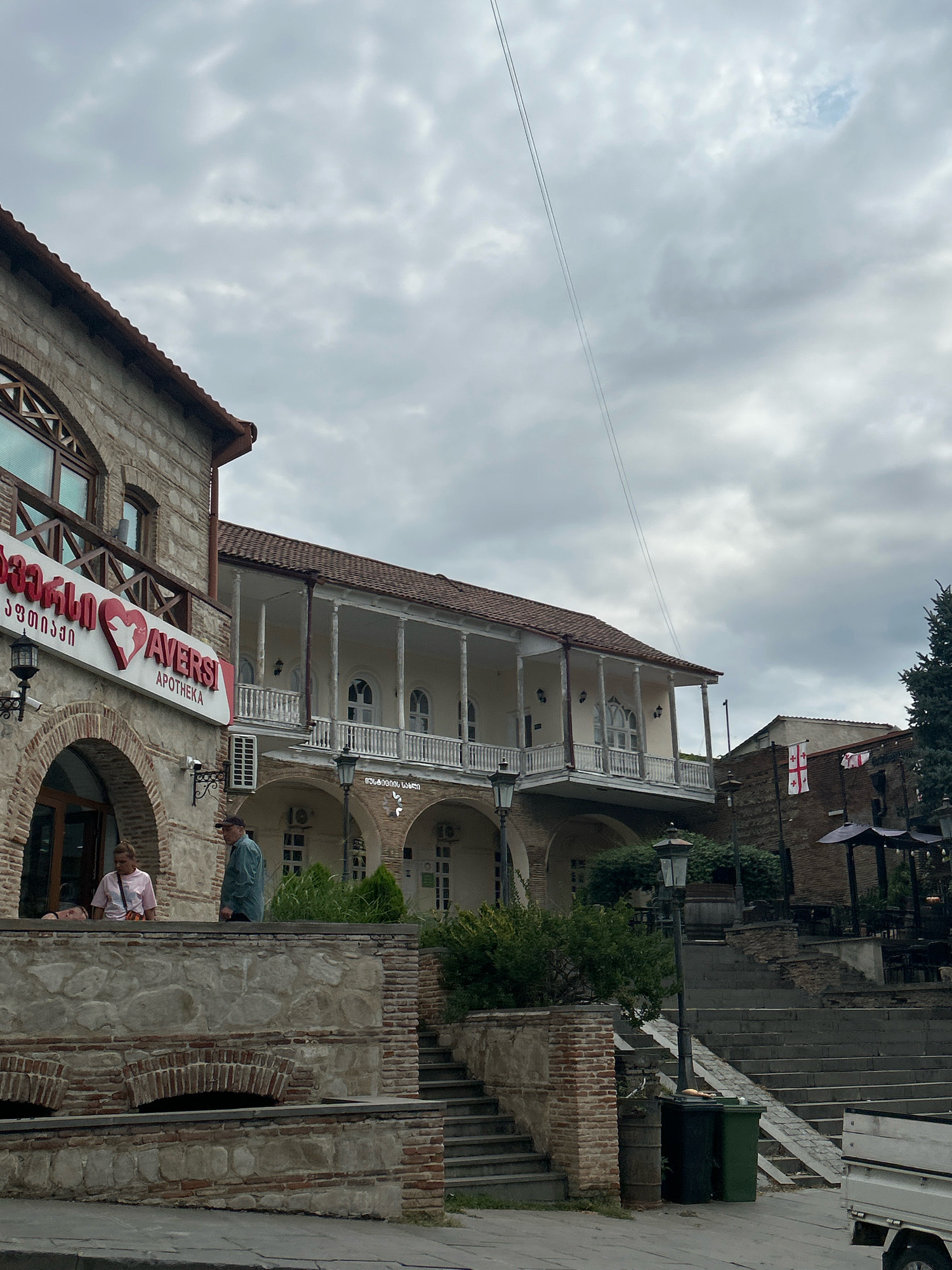 This image depicts a street scene in Sighnaghi, Georgia, a town known for its historic architecture. The foreground features a stone and brick building with arched entrances. A sign with red text and a heart logo, reading "AVERS APOTHEKA," is prominently displayed on the left side of the building. Two people are visible near the entrance of this building, one appearing to lean on a railing. To the right of this building, a large, multi-story structure with a long, covered balcony supported by columns dominates the midground. The balcony has white railings and arched windows. Further to the right, another building with brickwork is visible, adorned with Georgian flags. A steep set of outdoor stairs leads up to an area with outdoor seating and umbrellas. The environment is overcast, with a cloudy sky filling the upper portion of the frame. A utility line stretches diagonally across the sky. Two trash receptacles are positioned near the base of a lamppost on the right side of the image. A portion of a white truck is visible in the lower right corner.