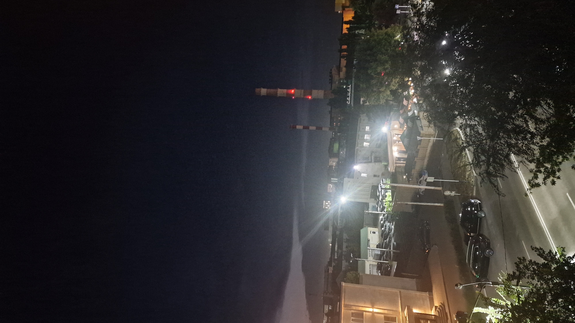 This image shows a nighttime view of a city, likely Kostanay, Kazakhstan, featuring industrial structures, buildings, cars, and a stormy sky.
In the distance, two tall, striped industrial smokestacks are visible. The taller one has red lights near its top. A dark, ominous cloud formation stretches across the sky in the upper left, with what appears to be lightning illuminating the clouds.
Closer to the foreground, several multi-story buildings are present, some with illuminated windows. Streetlights cast bright glows on the surroundings. A parking lot filled with cars is situated between buildings.
On the street in the lower part of the image, two black SUVs are parked. Another black car is visible on the road. A police car is parked near a fence. Trees with dense foliage are scattered throughout the scene, some obscuring parts of the buildings and street. The overall atmosphere is dim and somewhat moody due to the nighttime setting and the approaching storm.