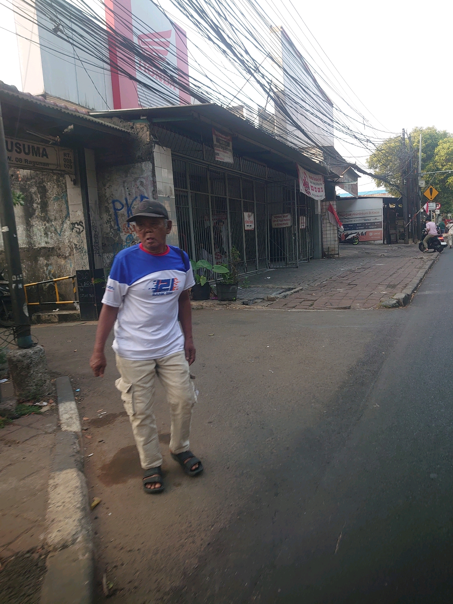 A man wearing a blue and white t-shirt, beige cargo pants, and black sandals walks on a paved sidewalk next to a road in South Jakarta, Indonesia. He is facing the camera and has a dark baseball cap on his head. Behind him, there is a building with various signs and awnings, including one with red and white lettering. The street is lined with power lines. In the background, there are trees and a building with a large red logo. A motorcycle is parked in front of a shop. The scene appears to be an urban environment during daylight.