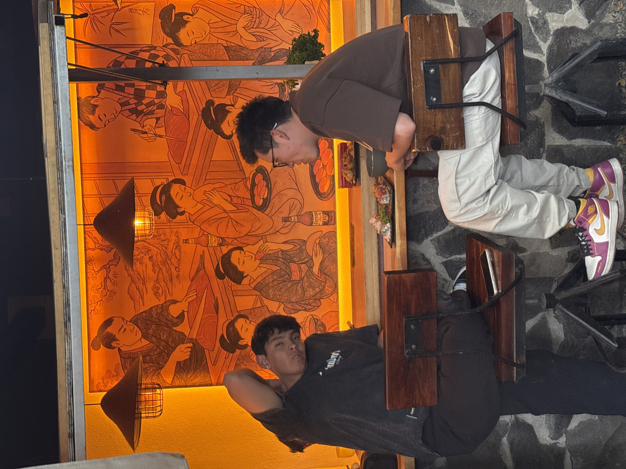 An indoor scene in a restaurant or bar located in Puebla, Mexico, depicts two young adult males seated at a high wooden counter. The male on the left, wearing a dark t-shirt and dark pants, is seated with his left arm behind his head, head tilted to the right, looking directly forward. An earring is visible in his left ear. A black smartphone rests on the seat of his dark wooden bar stool. The male on the right, wearing a brown t-shirt, light grey cargo pants, and multi-colored sneakers, is seated with his forearms resting on the counter, looking towards the left. He wears glasses. On the counter in front of him are plates containing sushi rolls and what appears to be fried food, along with a small bowl. The background consists of a large, illuminated mural with an orange glow, illustrating Japanese-style figures in traditional attire engaged in activities such as preparing food and eating; a bottle labeled "Corona" is depicted within the mural artwork. Two black, caged pendant lights hang above the counter. The wall beneath the counter is constructed of dark, irregular stone. A small green plant is visible on the counter to the right.