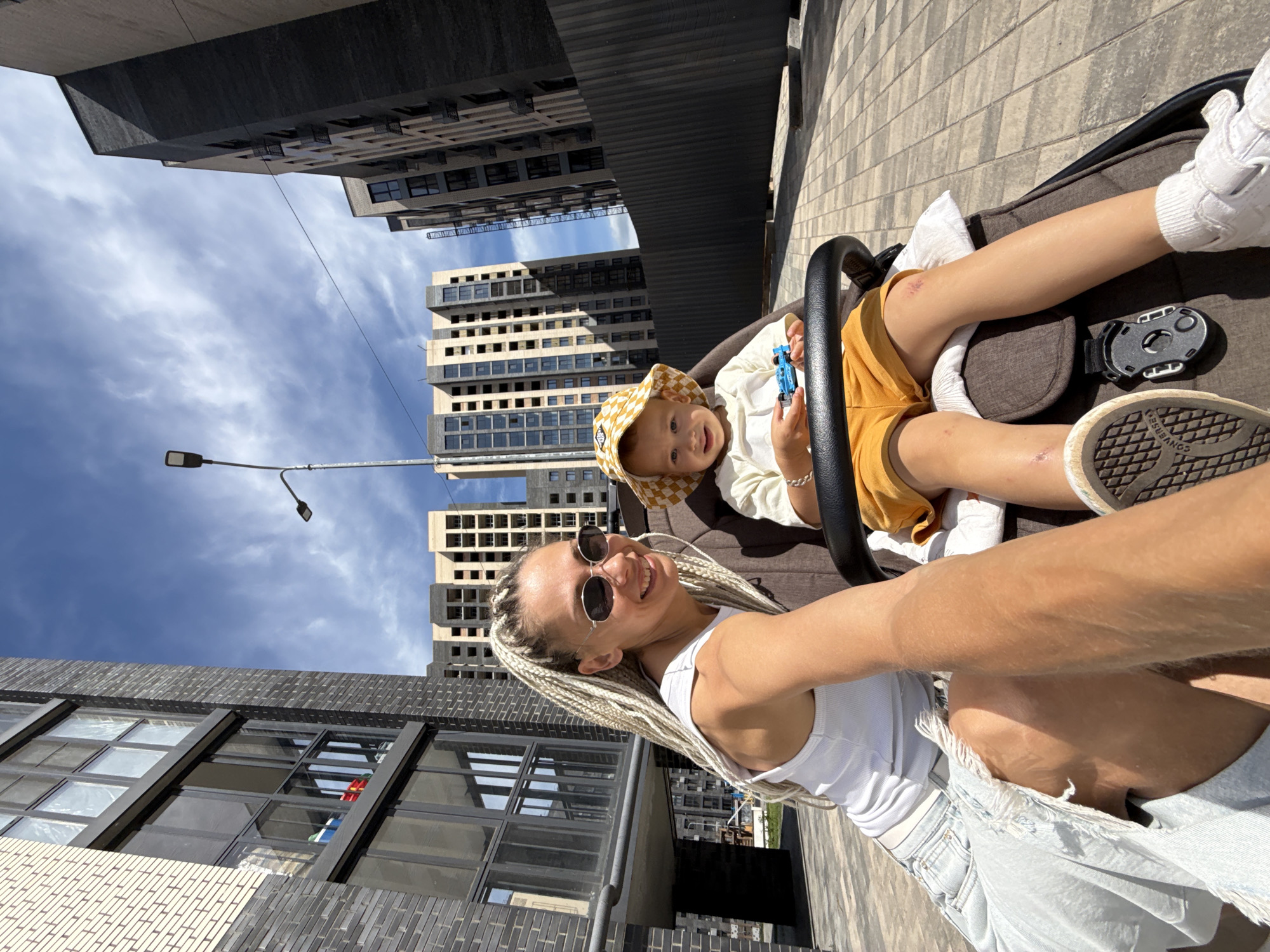 The image depicts a mother and her young child in a stroller, positioned outdoors in what appears to be a modern urban development area. The mother, with blonde braided hair and sunglasses, is smiling and looking towards the camera, holding the phone to take the selfie. She is wearing a white tank top and a light blue denim skirt. The child, wearing a yellow checkered bucket hat, a white t-shirt, and yellow shorts, is seated in the stroller, holding a small blue toy car.
The background features the facades of tall, contemporary apartment buildings under a clear blue sky with scattered clouds. One building is partially covered in light-colored brickwork and has large windows. Another is a dark-colored building with multiple stories and balconies. A dark wooden fence runs horizontally in the mid-ground. The ground is paved with interlocking grey and beige tiles. A street lamp is visible on the left side of the frame. The scene suggests a sunny day in an area that may be under construction or recently completed, based on the unfinished appearance of some buildings and the overall architectural style. The location is identified as Rusanovo, Russia.