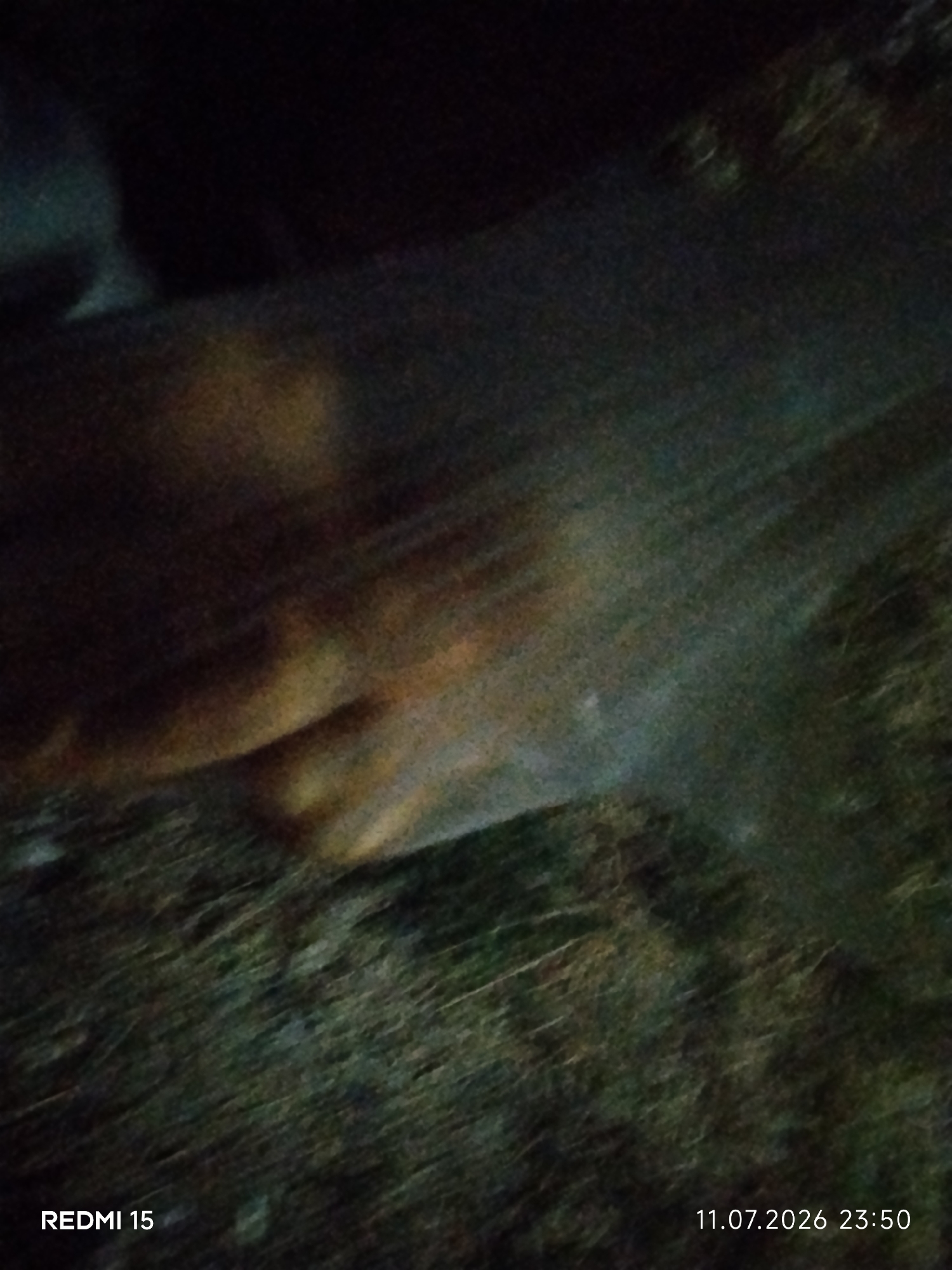 The image shows a close-up of a person's hand, possibly a foot, covered by a sheer, translucent fabric. The fabric has a shimmering quality, suggesting it might be made of a synthetic material or have metallic threads. The underlying skin tones are visible through the fabric, appearing warm and diffused. The background is dark and indistinct, with a textured, possibly grassy or earthy surface. The lighting is dim, with some highlights on the fabric and the skin. The image is overlaid with text indicating "REDMI 15" and a date and time stamp "11.07.2026 23:50". The scene appears to be indoors or outdoors at night, given the low light conditions and the date/time stamp. There is no specific indication of the location being Grinăuți, Moldova, as this information is not visually present in the image itself.