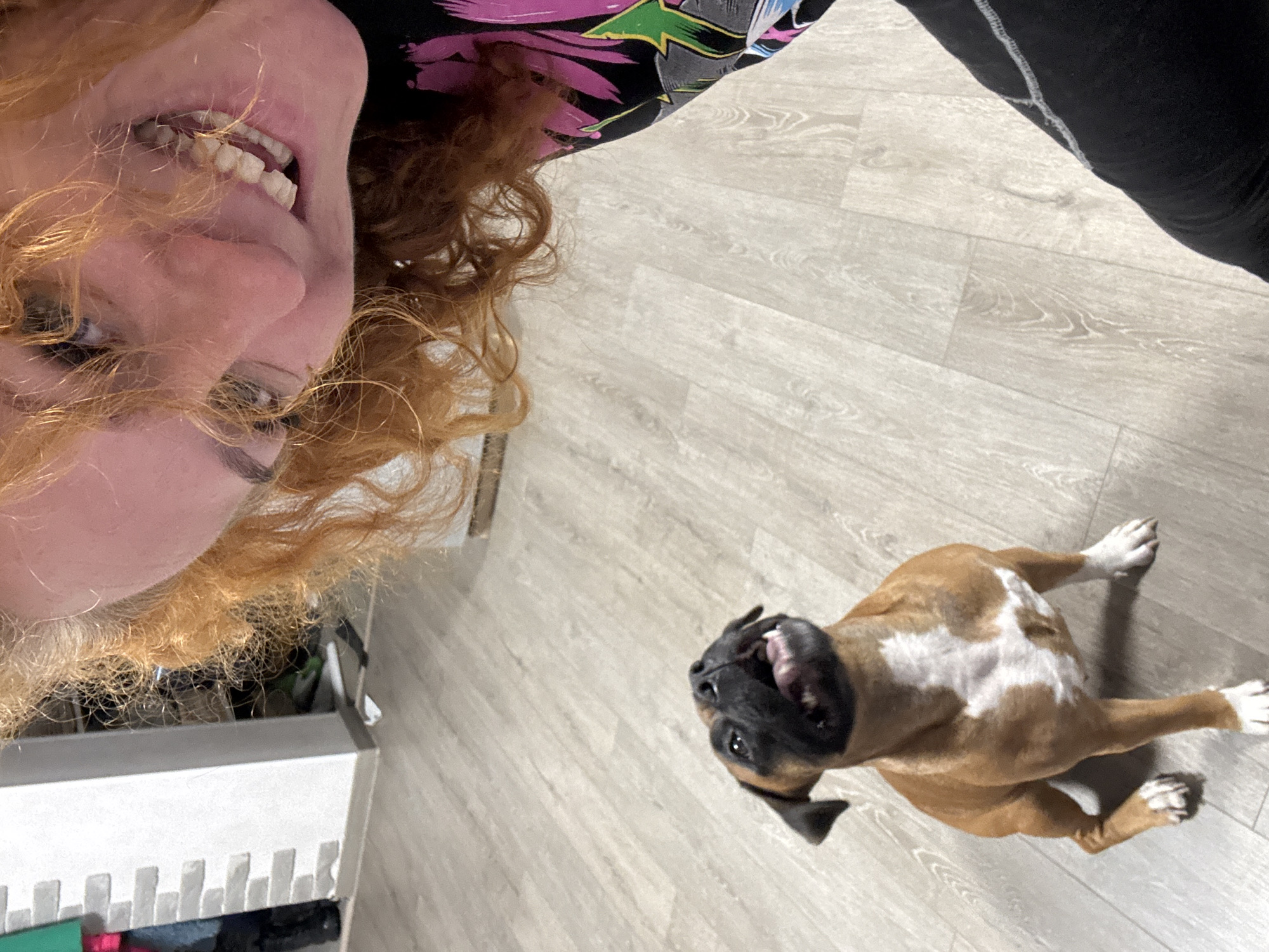 The image shows a woman with curly red hair and fair skin looking down at the camera. She is wearing a black shirt with a colorful floral pattern. To the right of her, on a light grey wood-look floor, is a brown and white boxer dog looking up at the camera with its mouth open. A white storage unit is visible in the lower left corner, and a pair of black leggings are visible in the upper right corner. The location is not identifiable as Chaikovsky, Russia from the visual information provided.
