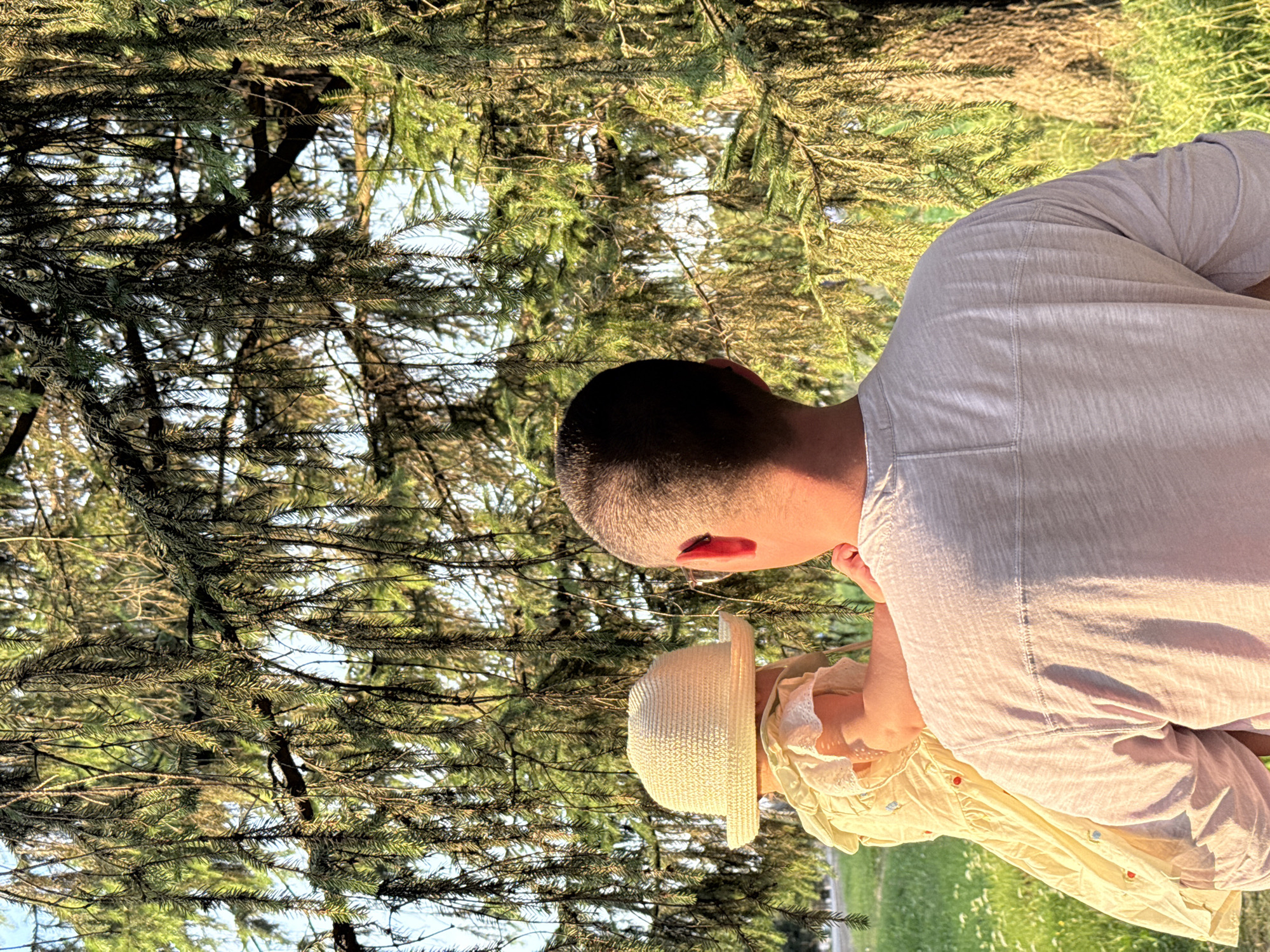 A person carries a baby in their arms, standing outside in a grassy area. The baby is wearing a light-colored hat and a yellow dress. The person holding the baby is wearing a light-colored shirt. They are positioned under the branches of a large evergreen tree, which fills much of the frame. The background shows a patch of grass with some small white flowers and sunlight filtering through the tree's needles. The scene appears to be in a park or natural setting.