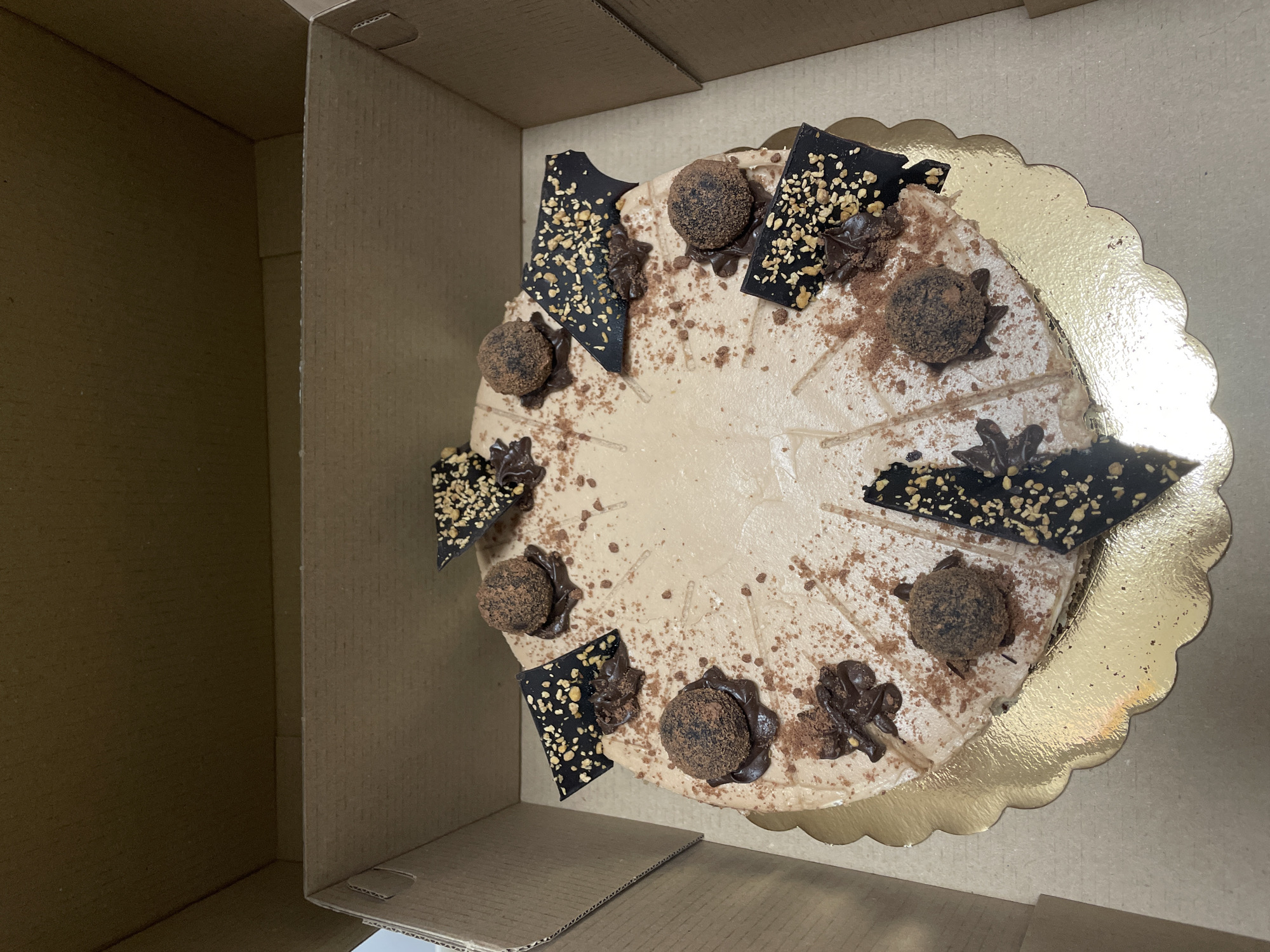 A top-down view of a chocolate cake in a brown cardboard box. The cake is decorated with chocolate truffles and dark chocolate pieces sprinkled with nuts. The cake rests on a scalloped, gold-colored base. The box is partially open, revealing the cake inside. The location is unknown, and there is no evidence to suggest the location is in Sofia, Bulgaria.