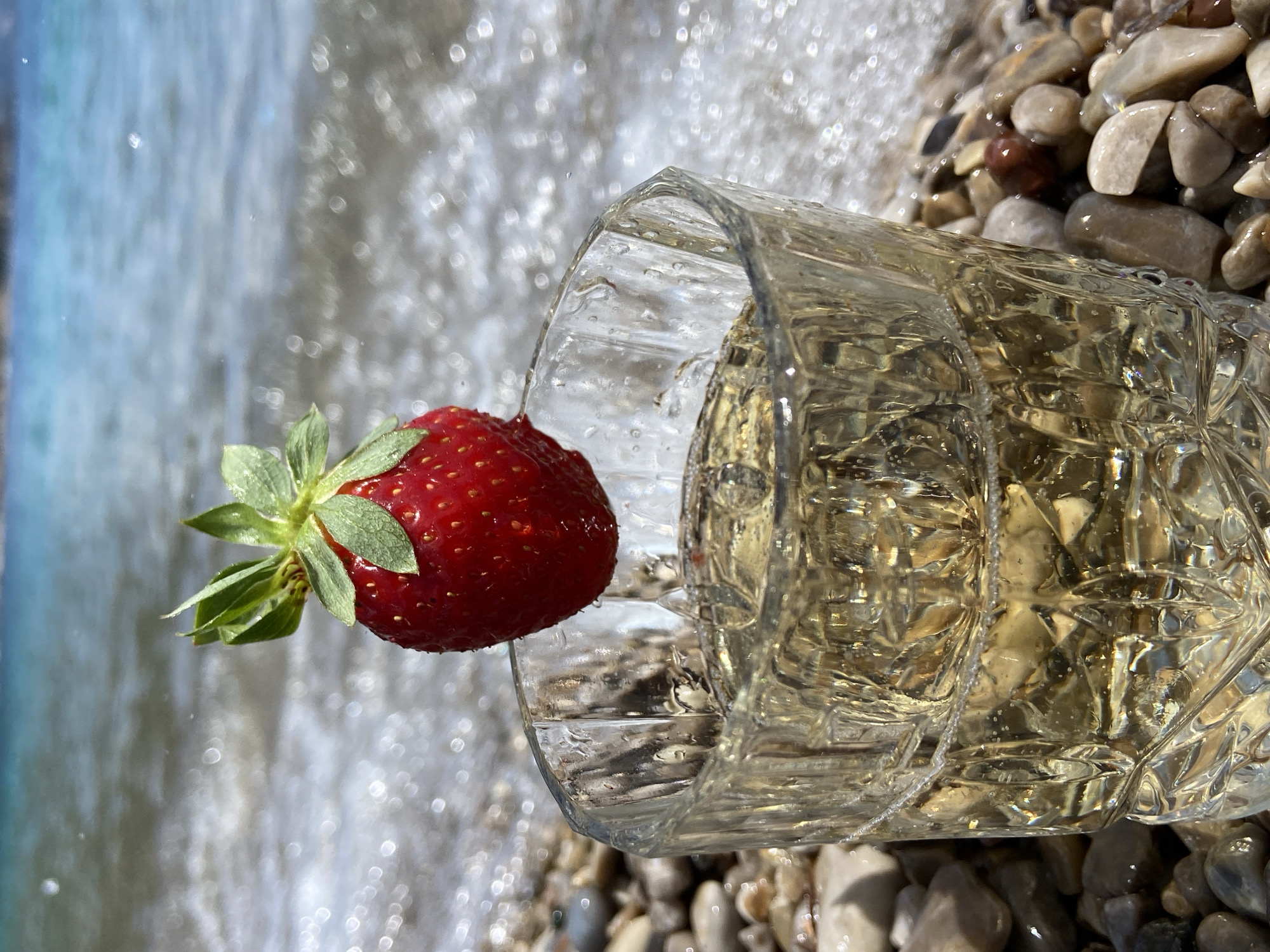 A clear glass with a beverage is lying on its side on a beach with small rocks. A strawberry is perched on the rim of the glass. The beach appears to be next to a body of water, possibly the sea. This scene takes place in Prijevor, Montenegro.