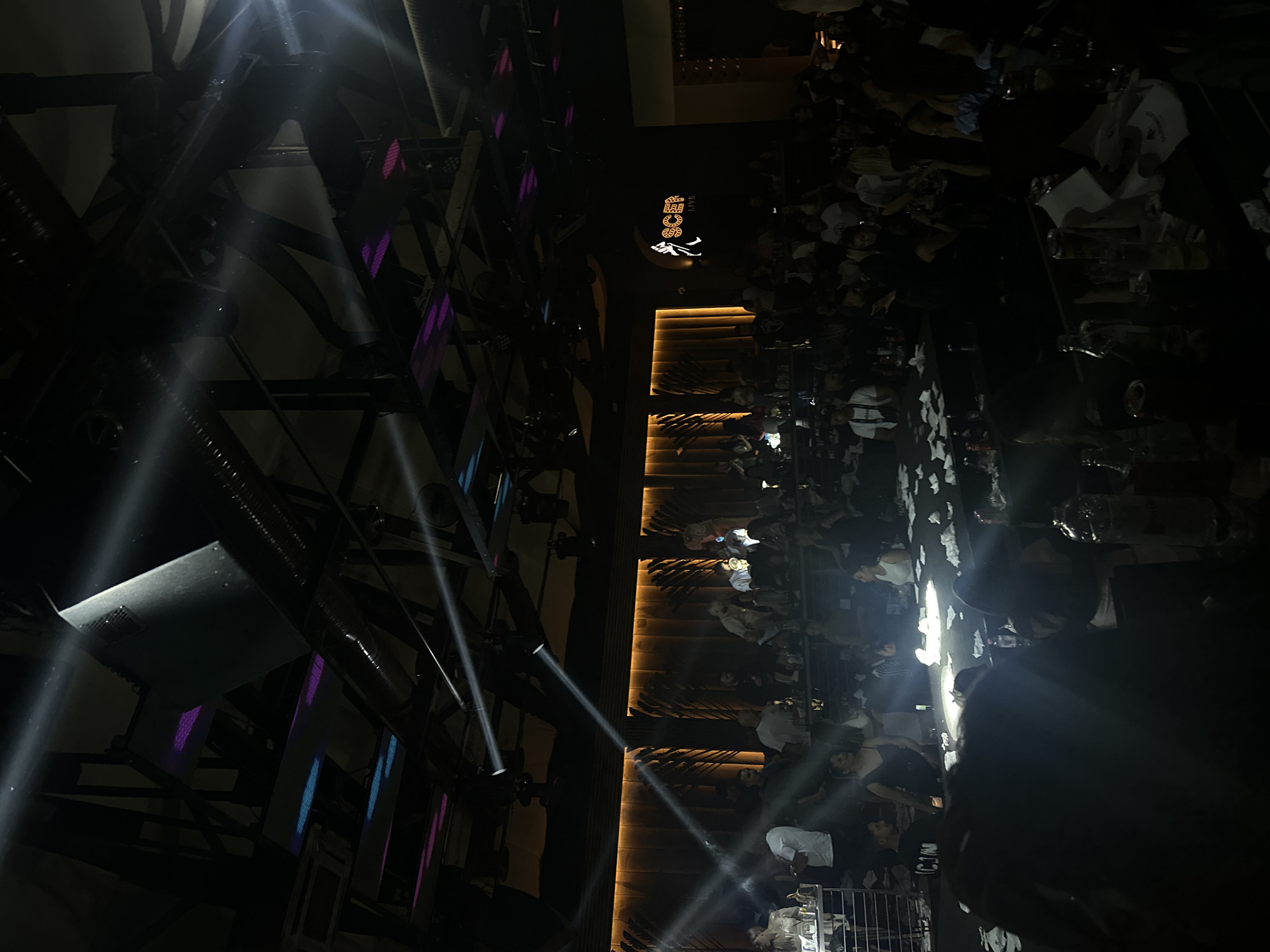 The image depicts an indoor venue, possibly a nightclub or entertainment space, with a dark, atmospheric ambiance. The ceiling is a complex structure of beams, pipes, and lighting fixtures, with spotlights casting strong beams of light downwards. Some of the lighting appears to be colored, with hints of purple and blue visible amongst the white beams.
Below the ceiling, the venue is populated by numerous people seated at tables. The tables are covered with what appear to be white napkins or serviettes, and bottles and glasses are visible on some of them, suggesting a bar or dining setting. The people are varied in their attire and are engaged in conversation or observing the surroundings. The overall lighting is dim, with the spotlights creating dramatic shafts of light that illuminate portions of the crowd and tables.
On the far wall, a prominent illuminated logo is visible. It features a silhouette of a walking figure and the text "SCEN LIVE" below it, indicating the name of the establishment. The back wall features a textured, possibly wooden, paneling with vertical, art-deco-style elements that are lit from behind, creating a warm glow.
The image captures a moment of activity within a dimly lit, social gathering space. The focus is on the interplay of light and shadow, the presence of a crowd, and the branded signage of the venue.