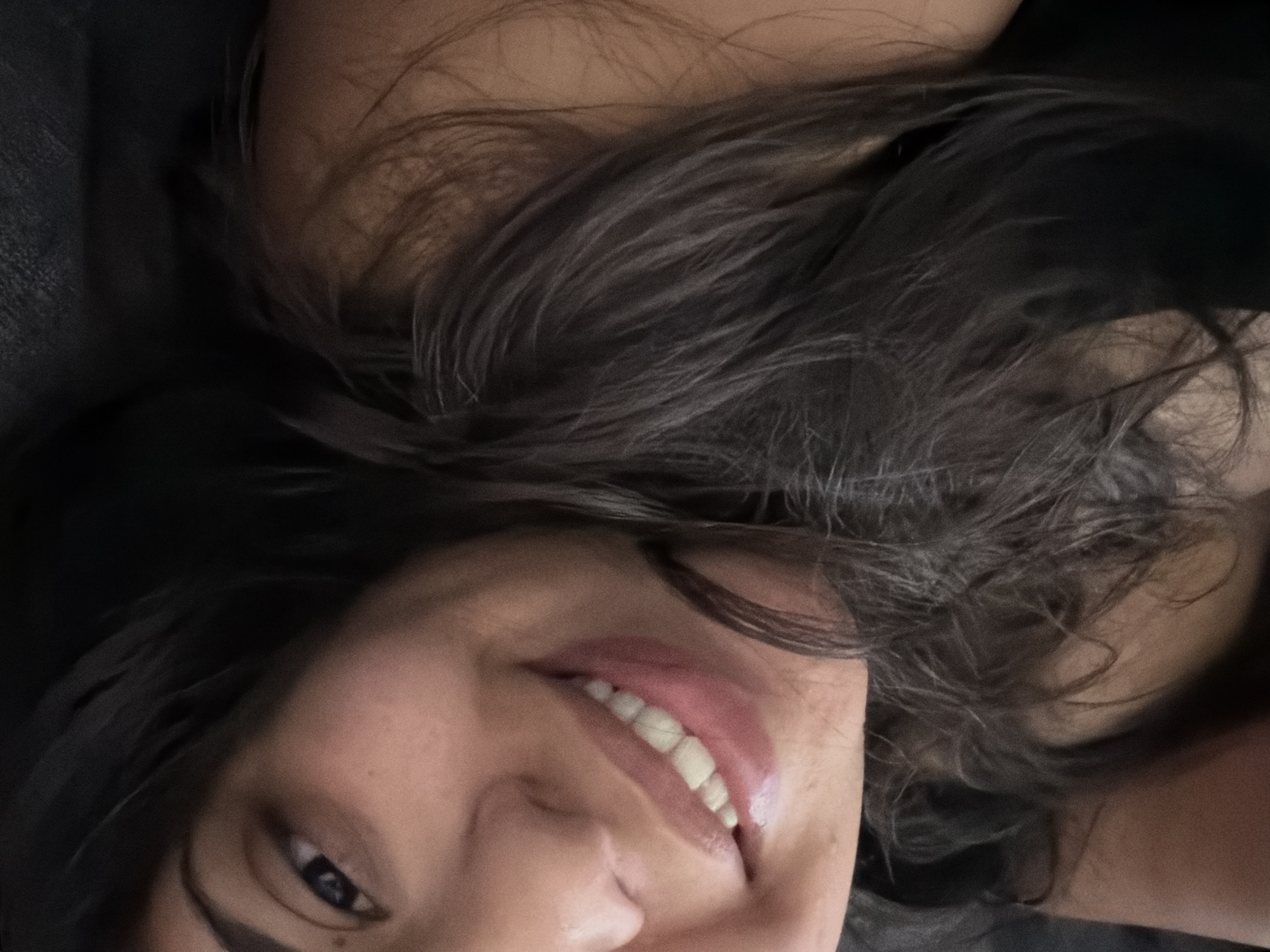 A close-up, high-angle selfie shows a young woman with dark hair smiling. She is wearing a dark top. The background is dark and indistinct. The image appears to have been taken indoors. There are no discernible features that would suggest a specific location in Duisburg, Germany.