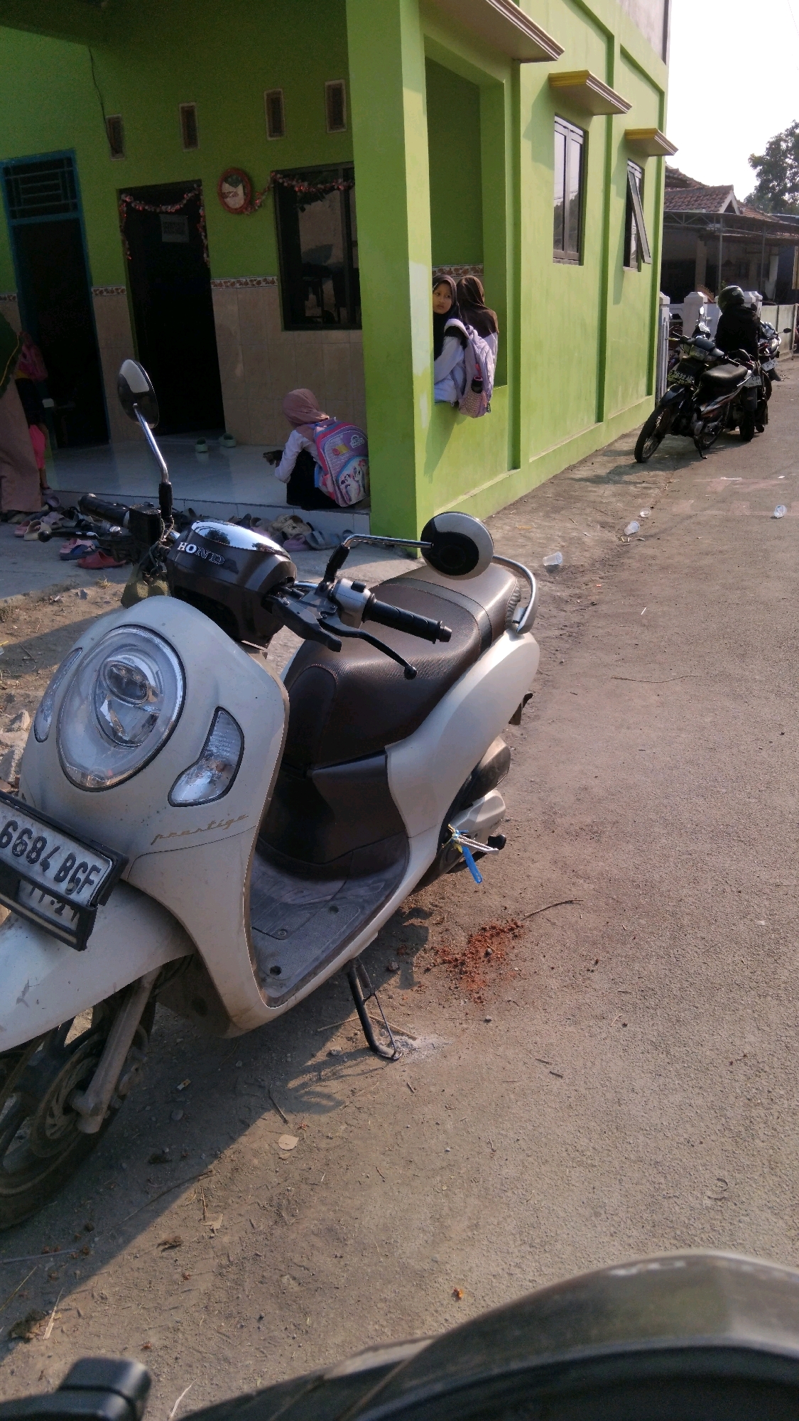 The image depicts an outdoor scene in front of a light green building, likely in Kalinyamat Wetan, Indonesia. A light-colored scooter with the license plate "6684 BGF" is parked on a paved surface. To the left of the scooter, a person in a brown hijab and pants is partially visible, standing near the entrance of the building. Inside an open window or alcove of the building, two young girls wearing hijabs are looking out. One girl has a light purple backpack, and the other has a pink backpack with a cartoon character. In the background, another scooter is parked to the right of the building, and a person wearing a dark helmet and jacket is seated on it. The environment appears to be a residential street with some debris and scattered plastic cups on the ground. The lighting suggests it is daytime, with shadows cast by the building and the scooter.