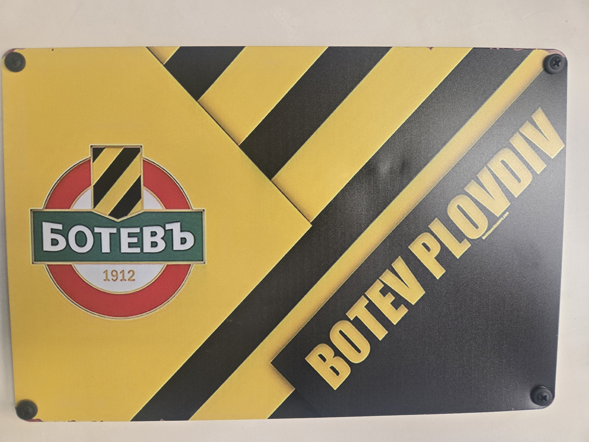 The image shows a rectangular sign with a yellow and black diagonal striped pattern. On the left side of the sign is a logo featuring the word "Ботевъ" in Cyrillic, written in white on a green horizontal banner. Below the word is "1912" in white, placed inside a red circle. On the right side, the words "BOTEV PLOVDIV" are diagonally written in yellow, against a black background. There are four black screws visible, one in each corner of the sign. The city is Plovdiv.