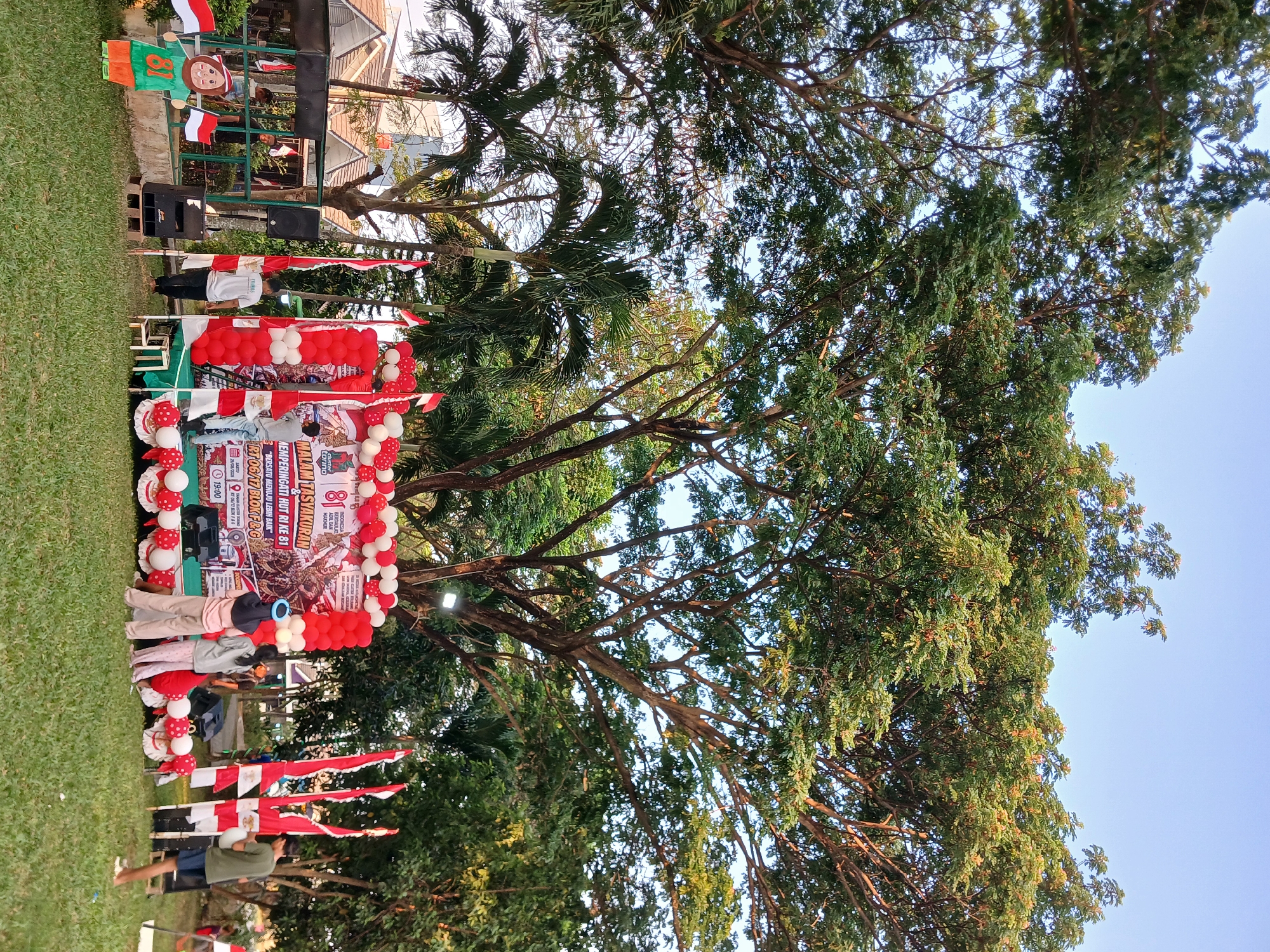 The image depicts an outdoor event likely in Kebalen, Indonesia, near a residential area with houses visible in the background. The scene is set under a large, mature tree with extensive foliage, suggesting a park or open green space. In the foreground, a stage is set up decorated with red and white balloons and flags, indicating a celebratory occasion, possibly a national holiday or anniversary, given the Indonesian flag colors. A banner on the stage reads "MALAM TASYAKURAN MEMPERINGATI HUT RI KE 81" (Gratitude Night Commemorating the 81st Anniversary of the Republic of Indonesia).
Several people are present at the event. On the stage, two individuals are attending to the setup, while a third is observing. In front of the stage, three women are interacting, one of whom appears to be wearing a blue headscarf. To the left of the stage, a cut-out figure of a child holding two Indonesian flags stands on the grass. Two large speakers are positioned on the ground near the stage, along with a small table. Indonesian flags on poles are prominently displayed around the stage area. The ground is covered in green grass, and the sky is clear blue, indicating it's daytime. The overall atmosphere suggests a community gathering or celebration.