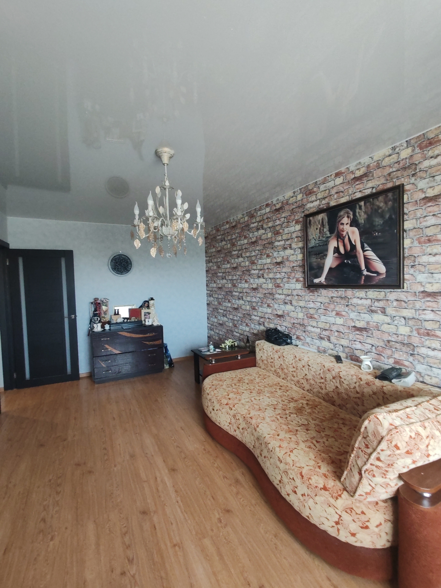 This image depicts an interior living space in the city of Petropavlovsk-Kamchatsky, Russia. The room features light brown laminate flooring throughout and a light-colored, possibly glossy, ceiling with visible reflections, including that of a light fixture or window and other ambient light sources. A multi-branched white and gold chandelier with candle-style lights and decorative hanging elements is suspended from the center-left of the ceiling. A circular vent or speaker grille is also visible on the ceiling to the left of the chandelier.
The right wall is covered with wallpaper designed to resemble an exposed brick wall, featuring a mosaic of brown, orange, and beige tones. A prominent, large, dark-framed picture is mounted on this brick-patterned wall, depicting a blonde woman in dark, form-fitting attire, kneeling in shallow water, looking towards the viewer.
A large, curved sofa upholstered in a patterned fabric of beige, orange, and brown hues is positioned along the brick wall. A black bag, a small white ceramic holder, a remote control, and other dark accessories are placed on the sofa cushions and backrest. Adjacent to the right armrest of the sofa is a small, dark wood side table with a lower shelf, holding a small bottle and another item on its upper surface.
The back wall, on the left side of the image, is painted a light blue or gray-blue color with a subtle textured pattern. A dark brown wooden dresser or chest of drawers with three drawers, adorned with lighter-colored decorative accents, rests against this wall. The dresser's surface displays an assortment of items, including bottles, small framed pictures, and decorative objects, along with a small mirror. Above the dresser, a circular wall clock with a dark frame and a decorative face is mounted. On the far left, a dark brown or black door with two vertical light-colored panels and a silver-colored handle is visible. A dark-colored bag is partially visible on the floor next to the dresser. No people are physically present in the scene, only depicted in the artwork.