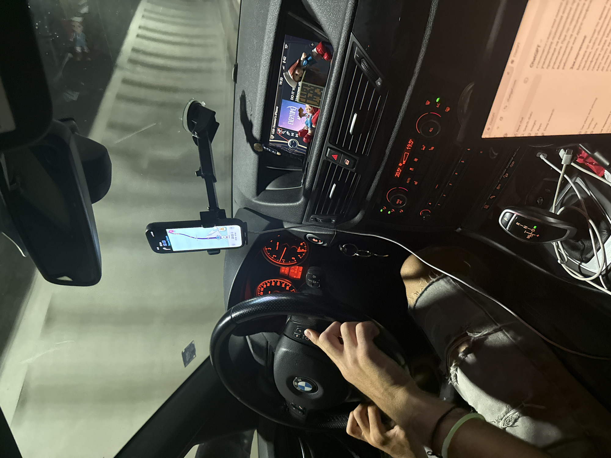 The image shows the interior of a car, likely a BMW, at night. The car appears to be in motion, as indicated by the blurred background of a tunnel. The driver's hands are on the steering wheel, and a smartphone is mounted on the windshield displaying navigation. The car's infotainment system is visible, showing music playback and a phone connection. A laptop is open on the passenger seat, displaying a webpage. The scene is consistent with a car traveling through a tunnel on a road near Dolna Vasilitsa, Bulgaria.