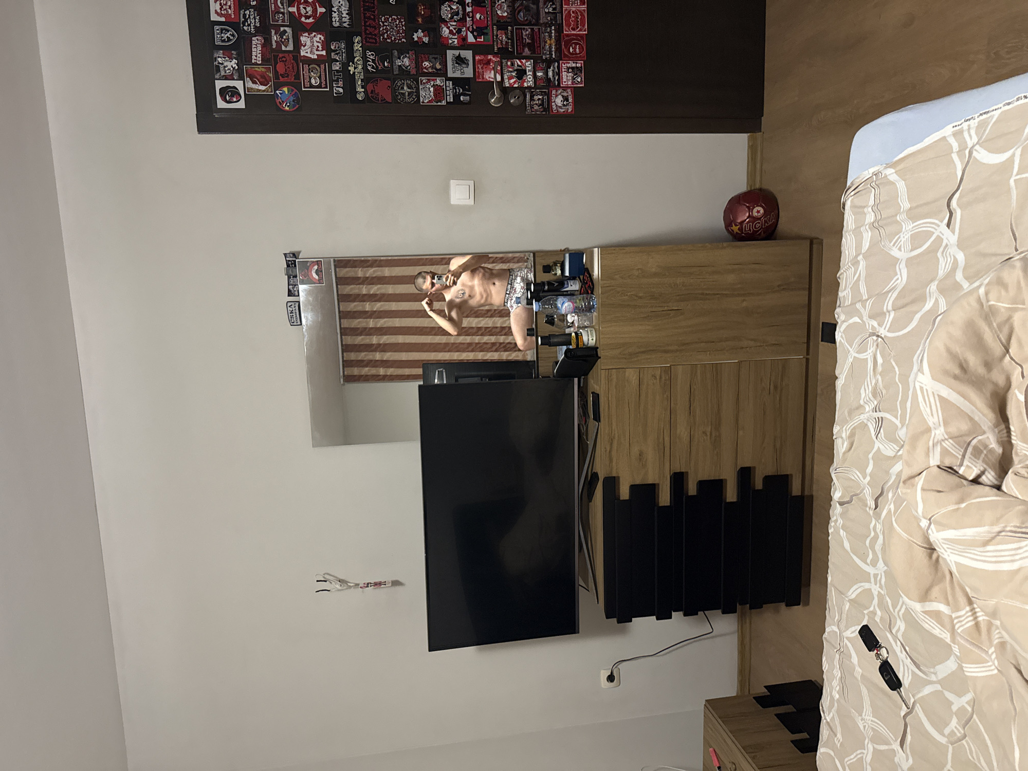 The image depicts the interior of a residential room, stated to be located in Blagoevgrad, Bulgaria. The room features light-colored walls and wooden laminate flooring.
A male individual is visible in the reflection of a large mirror, which is part of a wooden dresser unit. The man is shirtless, wearing patterned boxer briefs, and is captured taking a selfie with a smartphone held in his right hand while flexing his left bicep.
A large, dark-screen flat-panel television rests on the dresser, positioned to the left of the mirror. The dresser itself features a light wood finish on its main structure and dark, horizontal panels or drawers in its central section. Various personal items are arranged on the dresser to the right of the television, including several bottles (one appearing to contain water), a black wallet or small case, and a blue container.
To the right of the dresser, a dark brown door is heavily adorned with numerous stickers and patches, predominantly featuring insignia and text associated with CSKA Sofia football club and its supporter groups. Visible text includes "CSKA", "SOFIA", "ULTRAS", "OFFENDERS", "1948", "ARMY", and "ТРИБУНА СЕКТОР Г". A white electrical wall outlet is present on the adjacent wall near this door.
On the wall above the dresser, a white lanyard with a clip hangs. Two small "CSKA" stickers are applied to the wall above the mirror frame. An electrical outlet with a plugged-in black cable is located on the wall to the left of the dresser, near the floor.
In the foreground, a bed with a beige and white patterned duvet cover and a light blue fitted sheet is partially visible. A set of car keys with a black remote fob lies on the duvet. A red football, marked with a yellow star and the Cyrillic text "ЦСКА" (CSKA), is situated on the wooden floor in the bottom right corner, between the dresser and the door. A light wood side table is partially visible on the far left of the frame. The mirror's reflection behind the individual shows vertical striped window coverings in brown and beige tones.