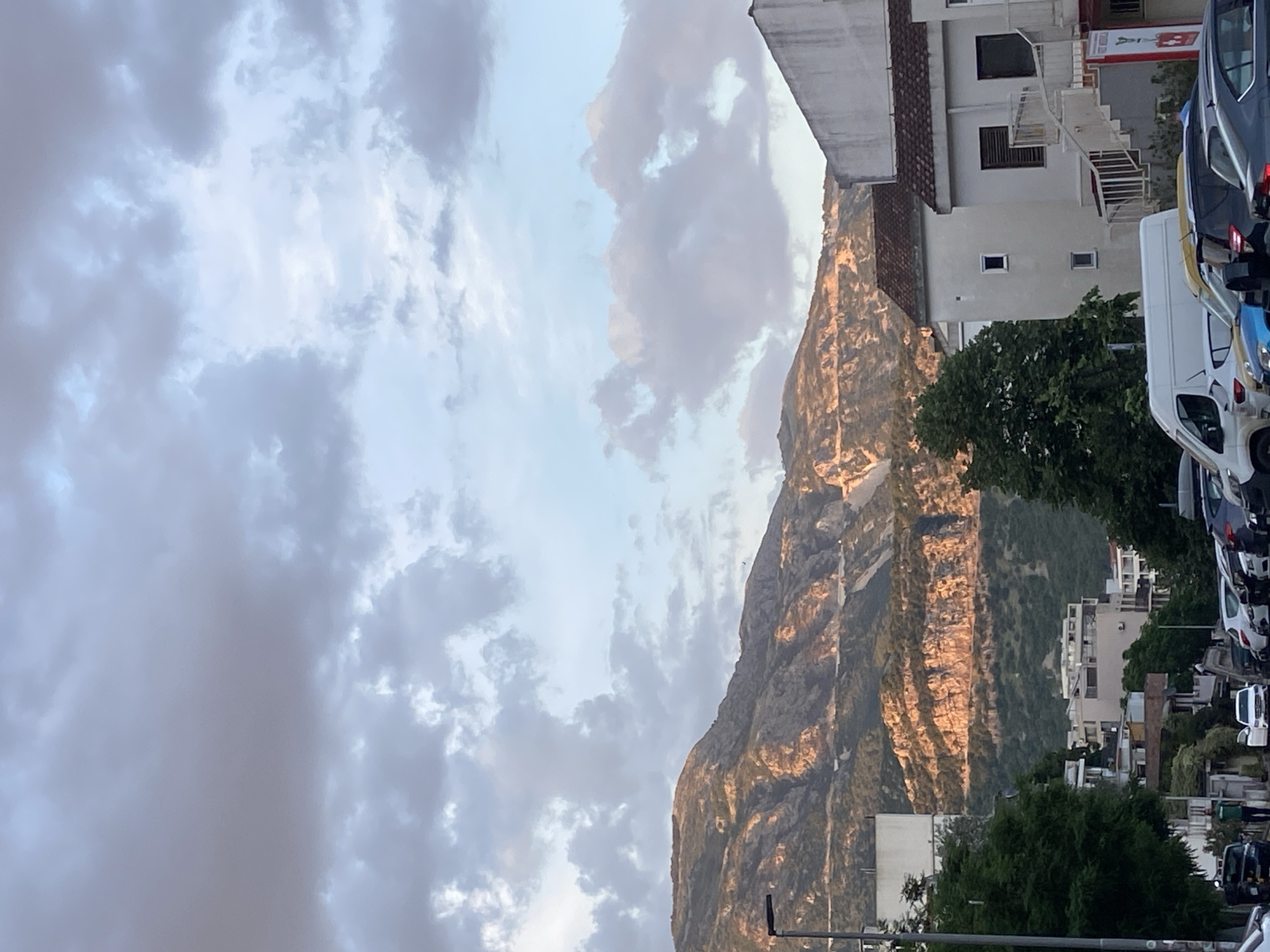 The image shows a view from a street in Budva, Montenegro. The shot is oriented vertically. The most prominent feature is a large, rocky mountain with visible striations. The mountain is illuminated by a warm, golden light, suggesting it is either sunrise or sunset.
To the left of the mountain, a sky is filled with clouds, some darker and grey, some lighter and appearing bluish-white. To the right of the mountain, there are several buildings and trees. Vehicles, including cars and vans, are parked along the street, indicating an urban setting. The tops of some streetlights are barely visible. The buildings are multi-story and appear to be residential or commercial. Overall, the scene combines natural and urban elements.