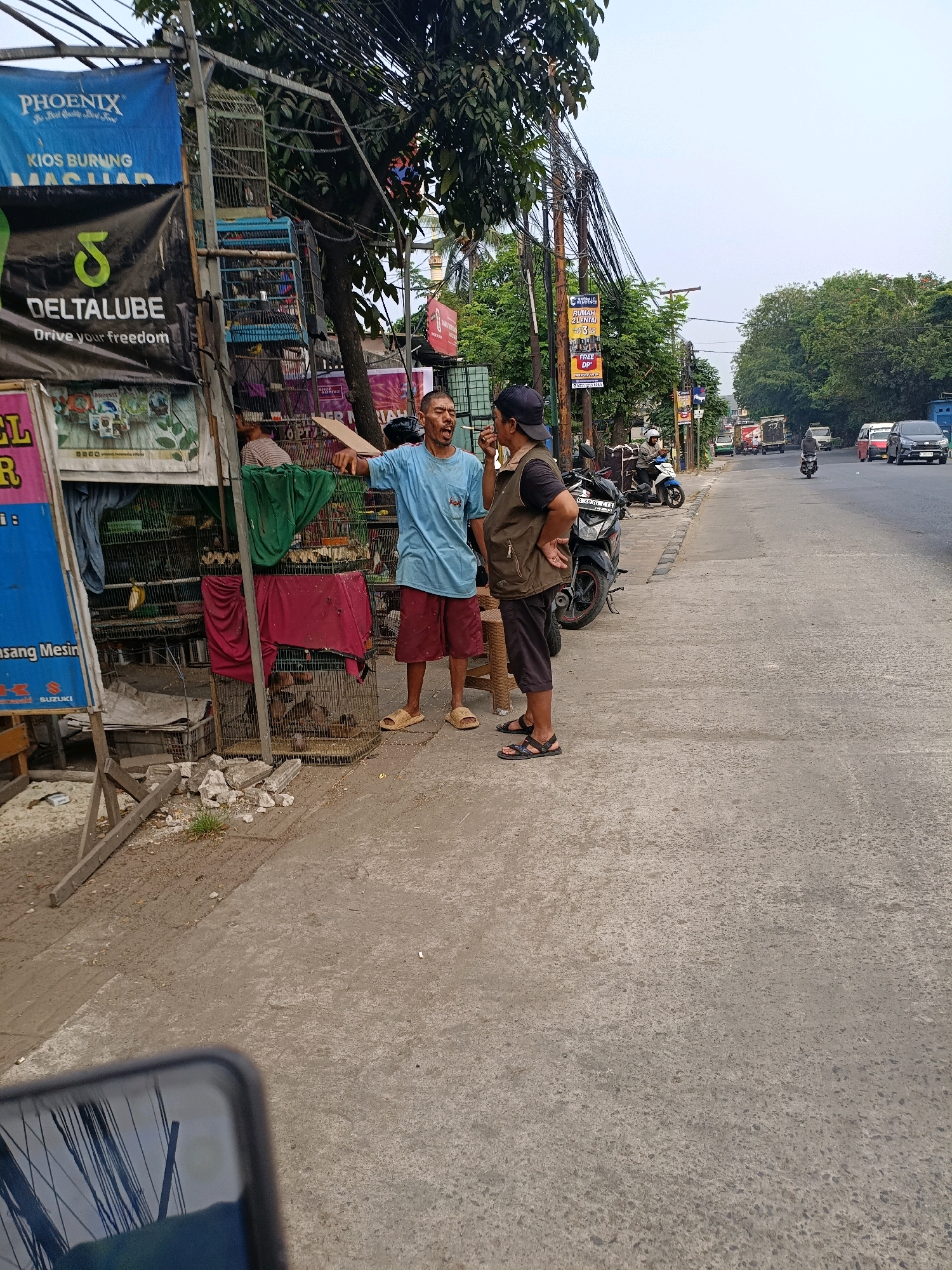 Two men are standing on a sidewalk next to a roadside stall selling birds. The man on the left is wearing a blue t-shirt and red shorts and is gesturing with his right hand while speaking. The man on the right is wearing a brown vest over a black shirt and dark shorts, with his hands on his hips. Behind them, several bird cages are stacked on a metal frame, with some birds visible inside. The stall has banners with various advertisements. The scene is set on a street in Tangerang, Indonesia, with a road, trees, and utility poles visible in the background, and traffic can be seen on the road. The overall environment appears to be an outdoor urban setting.
