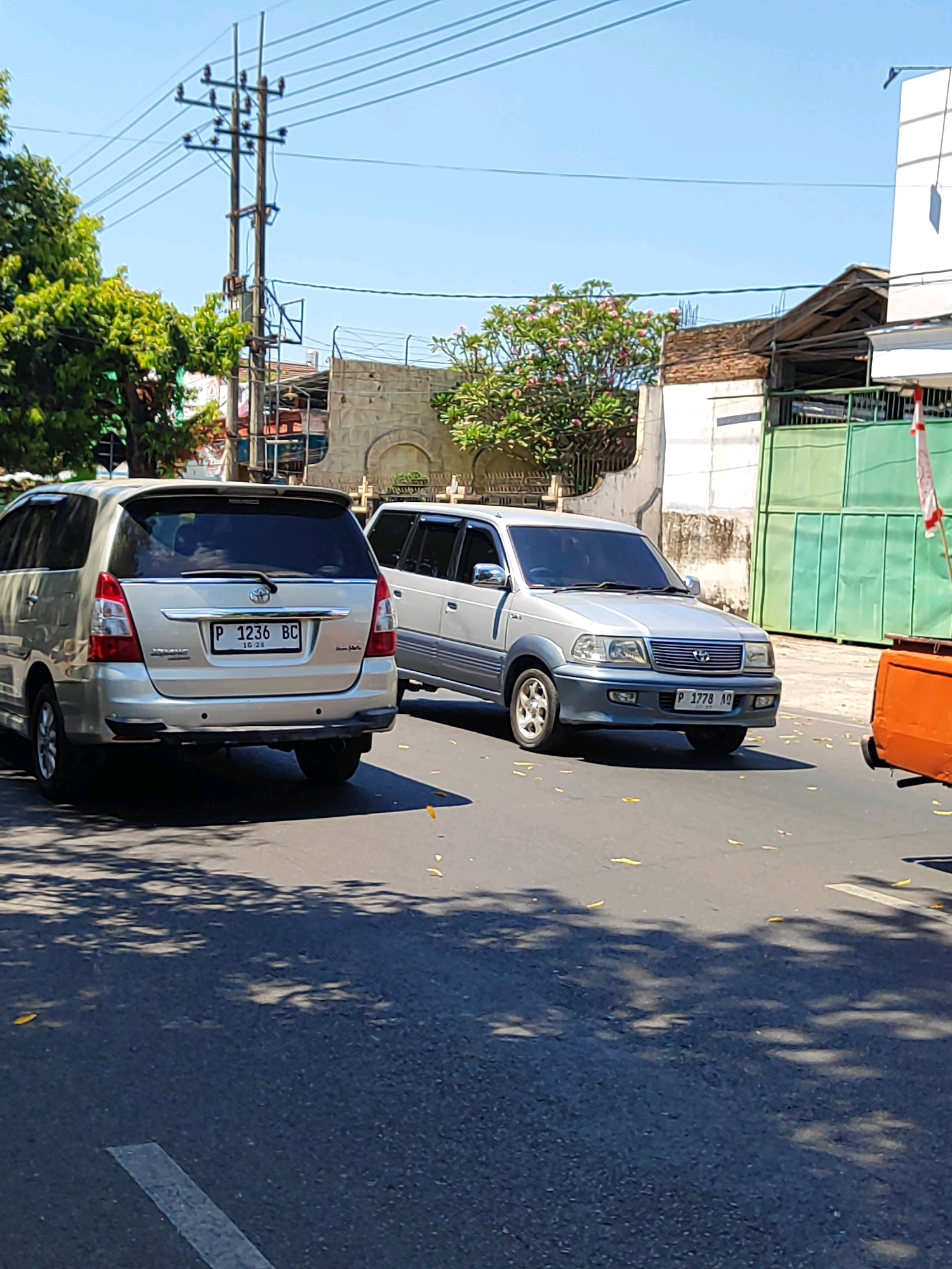 In Kotakulon, Indonesia, two silver SUVs are seen on a paved road under a clear blue sky. The vehicle on the left, a Toyota Innova with license plate P 1236 BC, is positioned closer to the viewer and is slightly angled to the right. The vehicle on the right, a Toyota Kijang with license plate P 1778 AQ, is further down the road and angled to the left. Trees with green foliage and some pink blossoms are visible on the left side of the road, as well as a power line pole with multiple wires. In the background, a building with a beige stone facade and arched windows is present, along with a green metal fence. The road surface shows scattered yellow leaves and shadows cast by trees. A partial view of an orange object, possibly a vehicle or equipment, is visible on the far right of the image.