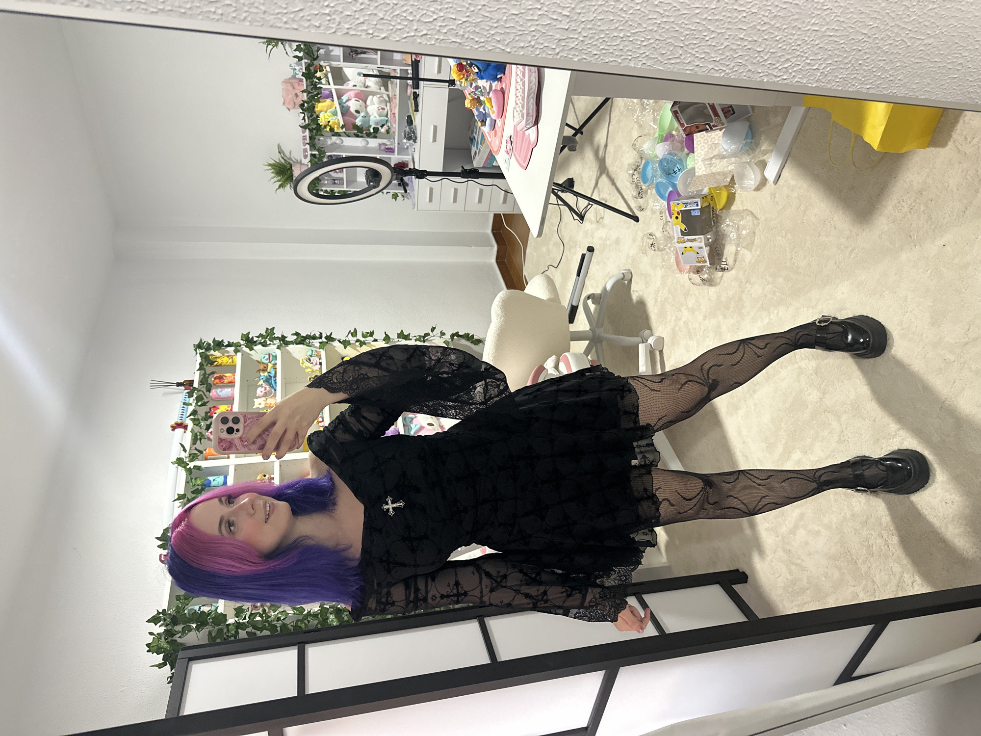 In what appears to be a bedroom or personal studio, a young woman with purple and pink dyed hair is taking a mirror selfie. She is wearing a black off-the-shoulder dress with lace sleeves and a cross pendant. Her legs are covered in black patterned tights with a spiderweb design, and she is wearing black platform shoes. The room is decorated with plants, shelves filled with figurines, and a desk with a ring light. The overall impression is one of a creatively decorated personal space, possibly used for content creation. There is no information in the image to suggest a location in Oviedo/uviéu, Spain.