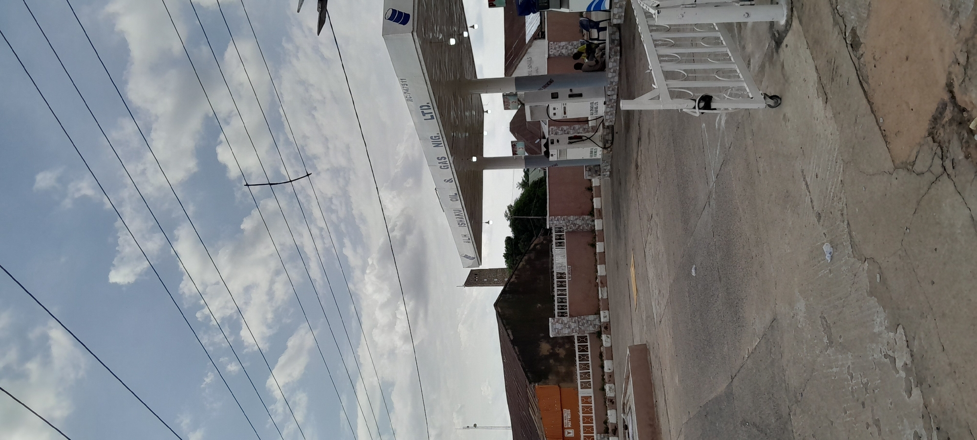 The image shows a gas station in Jalingo, Nigeria. The gas station features a white canopy with the text "ALH ISHAKU OIL & GAS NIG. LTD." printed on it. Several fuel pumps are visible under the canopy, with two individuals seated on chairs nearby. Behind the gas station, a building with a rusty roof is visible, and further in the background, a tall, slender tower can be seen. The foreground is a concrete paved area, with some debris on the ground, and a white metal gate with wheels is positioned in the lower right corner. Overhead, several power lines crisscross the sky, which is partly cloudy.