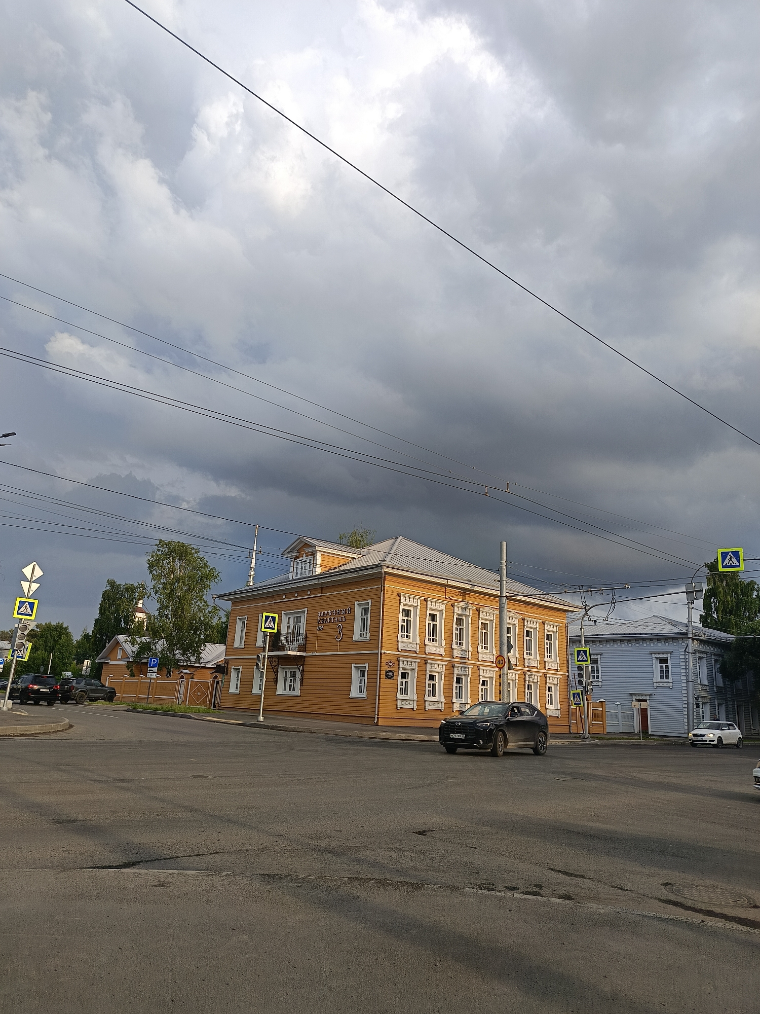 The image shows a street intersection with several buildings and cars. In the foreground, the asphalt road is visible with white crosswalk markings. A black SUV is driving through the intersection from the left side of the frame.
On the right side of the intersection, a two-story orange wooden building with white trim and numerous windows stands prominently. A pedestrian crossing sign is placed in front of it. Adjacent to the orange building is a light blue two-story wooden building. Another pedestrian crossing sign is visible in front of this building. A white car is parked on the road in front of the blue building.
In the background, more wooden buildings and trees are visible. The sky is overcast with dark, dramatic clouds, suggesting an impending storm. Electrical wires are strung across the sky, supported by poles. There are also traffic lights and street signs present at the intersection. The scene appears to be in an urban environment, likely a town or city, but the specific location is not identified.