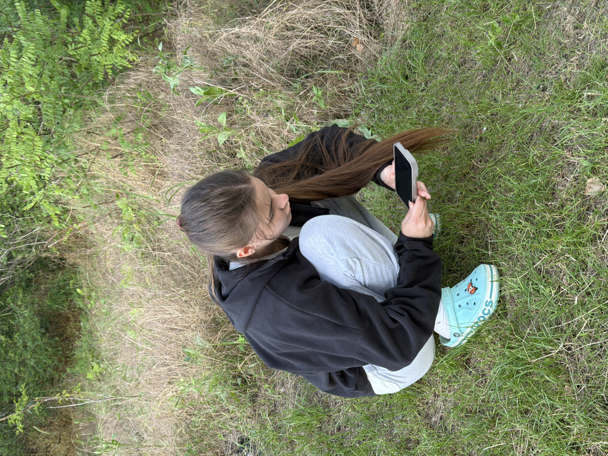 A young female is crouching on grass in an outdoor environment. She is wearing a black hoodie, light grey sweatpants, and light blue Crocs with a graphic on the side. She is holding a smartphone in her hands, looking down at it. The background consists of dry grass and green foliage, suggesting a natural or semi-natural setting, possibly a park or a less-maintained area within Obukhivka, Ukraine.