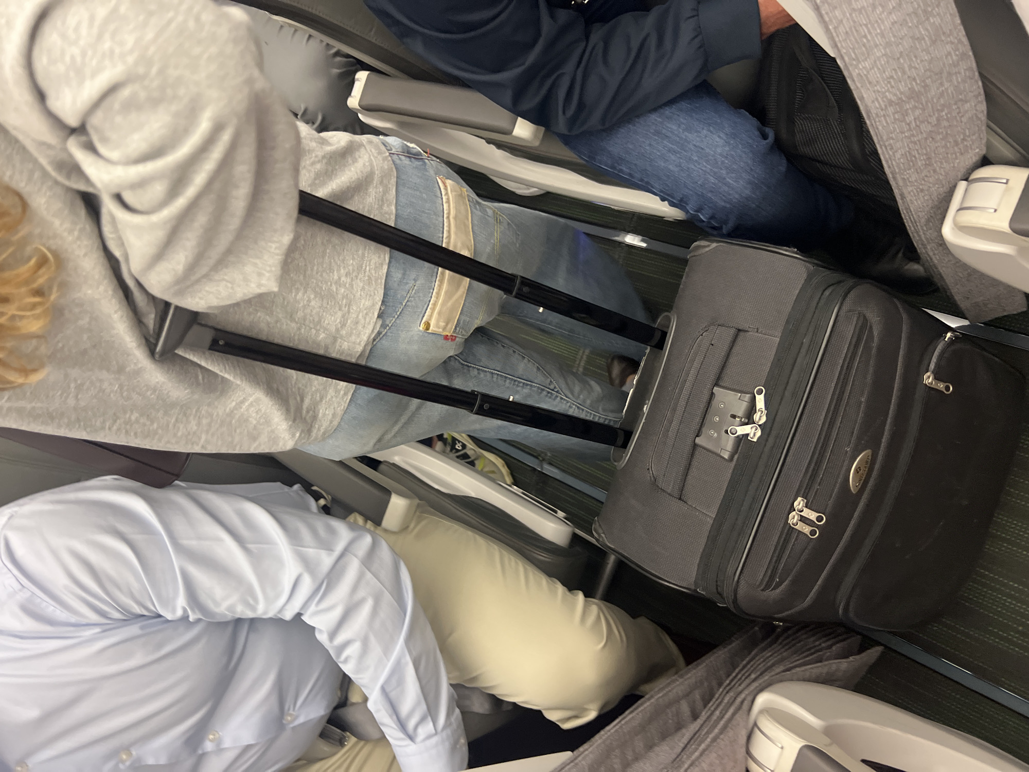 The image captures a close-up view of passengers seated in an airplane cabin, with their luggage positioned between seats. On the left, a person wearing a light blue button-down shirt is partially visible, their arm resting on their lap. Next to them, a person in a grey hoodie and blue jeans is seen from the waist down, holding onto the handle of a black rolling suitcase. This suitcase is directly in front of them, taking up a significant portion of the frame. To the right, another passenger's legs, clad in blue jeans, are visible, along with a dark jacket. The floor of the cabin appears to be covered in a dark carpet with faint green stripes. The overall scene suggests a typical situation during air travel, with passengers and their belongings arranged in a confined space. The location of Moreira, Portugal, is not discernible from the visual information provided in the image.