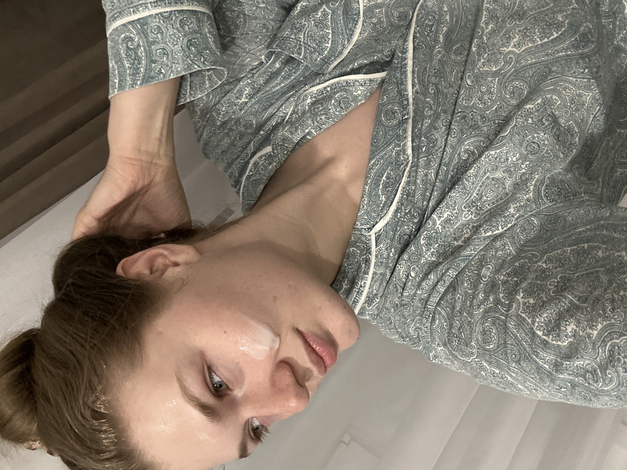 An indoor scene depicts a female individual, appearing to be a young adult, captured from the chest up within a private residence in Kazan, Russia. Her light brown hair is gathered into a high bun; some frontal strands exhibit a damp or treated appearance. Her eyes, light in color, are cast downwards and left. A white, creamy substance is visible on the subject's right cheek, extending from below the eye to the jawline. She wears a pajama top featuring a teal and white paisley pattern with white piping. The subject's right hand is raised to the back of her head, fingers contacting her hair. The background is an indistinct light-colored surface, with a dark, blurred element visible in the upper right quadrant.