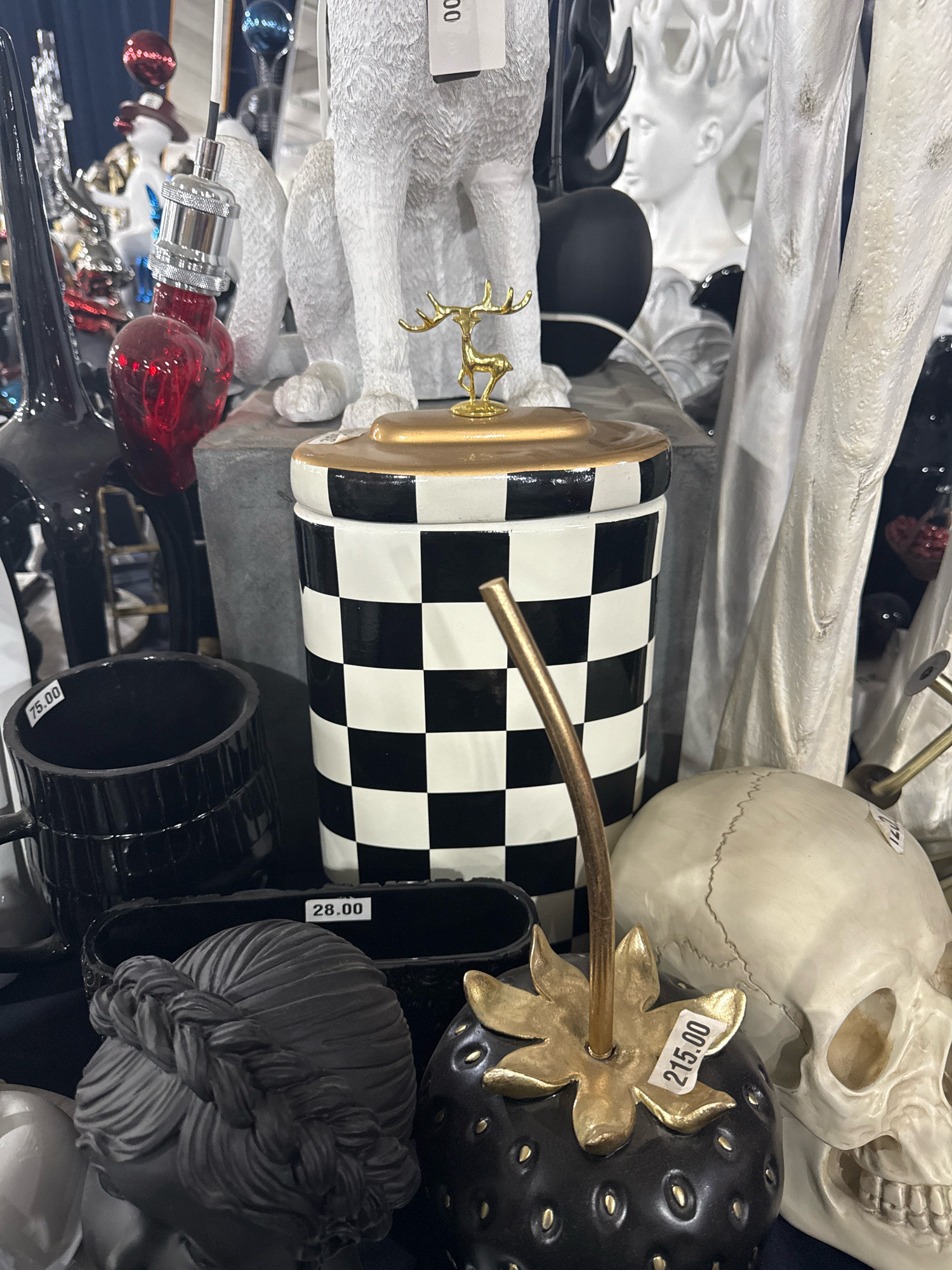 This image shows a display of decorative items in what appears to be a shop or market. The foreground features a black ceramic strawberry with a gold stem and leaves, a black bust with braided hair, a black mug with a "75.00" price tag, and a white skull. Behind these, a black and white checkered jar with a gold reindeer figurine on top is prominent. Further back, there are various other decorative objects, including a white dog statue and a white bust of a person. The overall setting is indoor, with some lighting visible. There is no indication of the city of Tbilisi, Georgia, in the image.