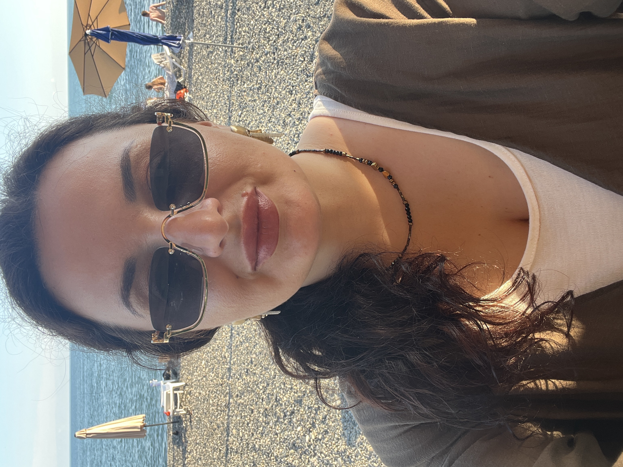 A woman with dark hair and sunglasses is taking a selfie on a pebble beach in Sokhumi, Abkhazia. She is wearing a white tank top and a brown cardigan. In the background, the Black Sea is visible, along with beach umbrellas, lounge chairs, and a few other people. The sun is shining, casting shadows on the beach.