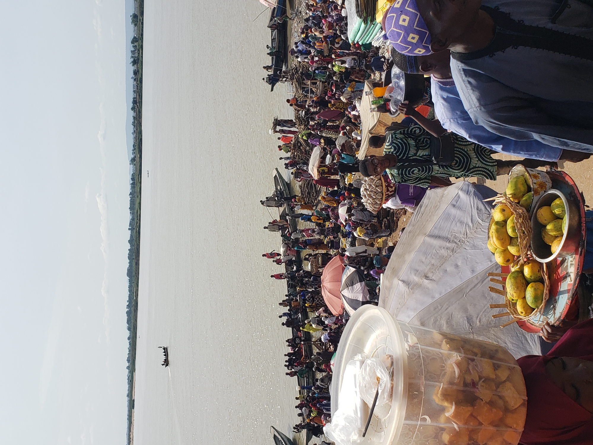 The image depicts a bustling scene at the confluence of the Niger and Benue Rivers in Lokoja, Nigeria. A large crowd of people has gathered on the shoreline. Boats are docked along the water's edge. The crowd is densely packed, with individuals carrying goods and wares. Several individuals are selling fruits (mangoes) and cooked food (puff-puff) from trays. There are white canopies and colorful umbrellas among the crowd. The environment is dusty with sparse vegetation. The river extends to the horizon, meeting a light blue sky. A small boat with passengers is seen on the water.
