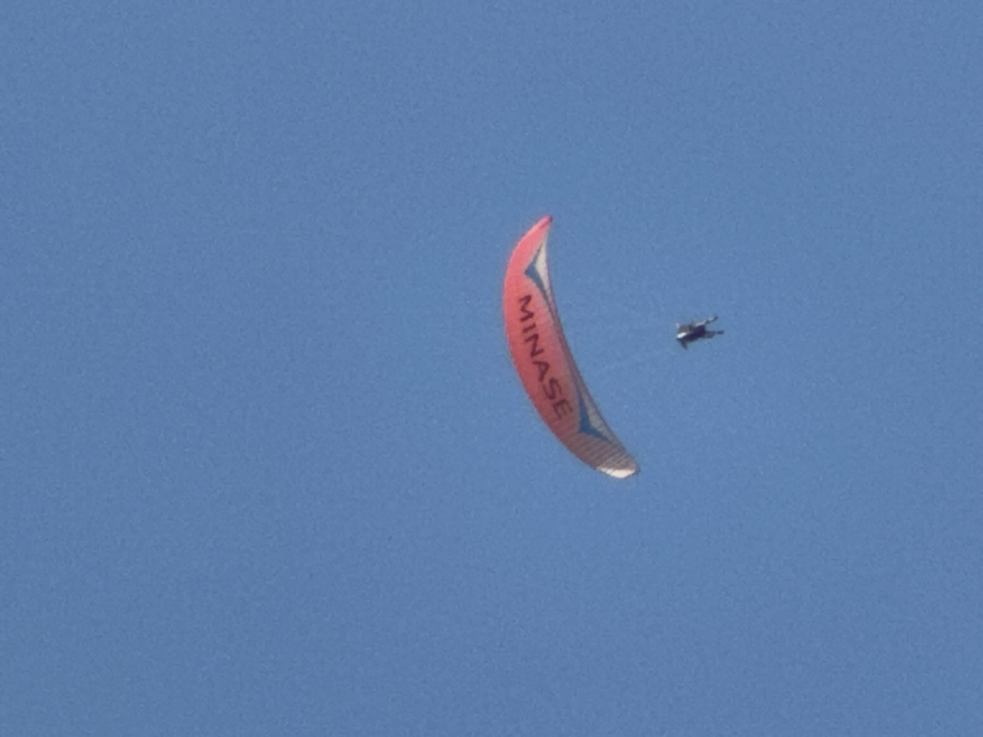 A paraglider, with the word "MINASE" visible on their wing, is soaring through a clear blue sky. The paraglider appears to be a single person, suspended beneath the colorful wing. The wing is primarily red and white, with a blue accent stripe along its edge. The paraglider themselves is a dark silhouette against the bright sky, making it difficult to discern specific features. The scene is set in Boreti, Montenegro, and the bright sky suggests it is daytime, likely with good weather for flying. The image captures a moment of freedom and adventure as the individual glides through the air, enjoying the expansive view.