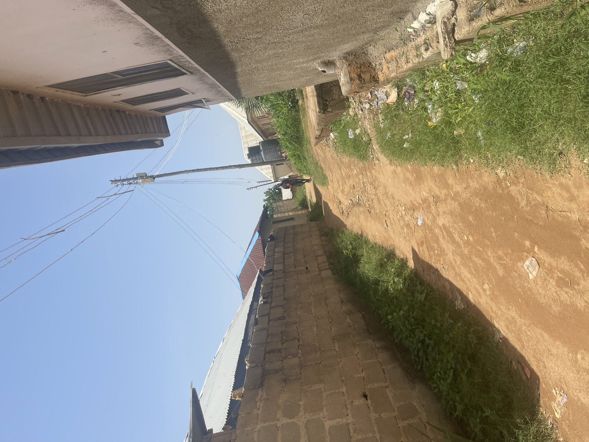 This media file depicts an outdoor scene in Rafin Par, Nigeria. The image is oriented vertically, with the sky taking up a significant portion of the frame. The foreground features a dirt path lined with overgrown green grass on the right and a brick wall on the left. Scattered amongst the grass and along the path are pieces of litter, including plastic bags and wrappers, indicating a lack of proper waste disposal.
In the background, several structures are visible. On the left, a building with a light-colored facade and rectangular windows can be seen. To the right of this building and further down the path, there are more structures, including one with a corrugated metal roof and another made of brick. Two large black water tanks are also prominent features on top of one of the buildings. A telephone pole with numerous wires stretches across the middle of the image.
A person is visible on a bicycle further down the dirt path, suggesting some form of movement or daily activity is taking place. The clear blue sky suggests it is daytime, and the lighting indicates sunny weather. The overall impression is one of an informal settlement or rural area, with a mix of functional infrastructure and signs of environmental neglect. There is no visible text in the image.