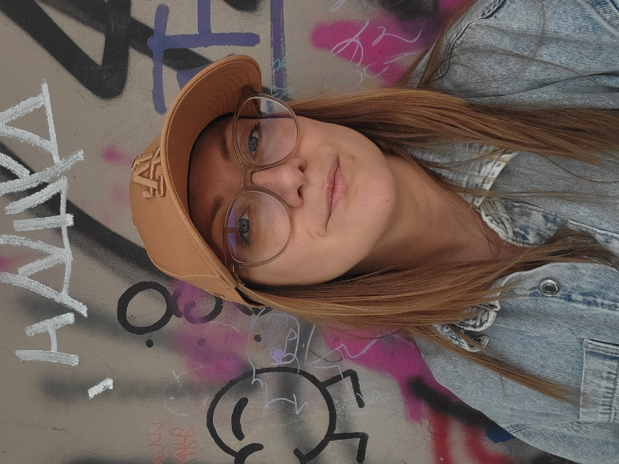 A young woman with long brown hair, wearing a tan baseball cap with the letters "LA" and round glasses, is posing against a graffiti-covered wall. She is wearing a light-wash denim jacket. The wall has various graffiti markings in white, black, purple, and pink. There is no specific information available to determine the precise location in Budva, Montenegro.