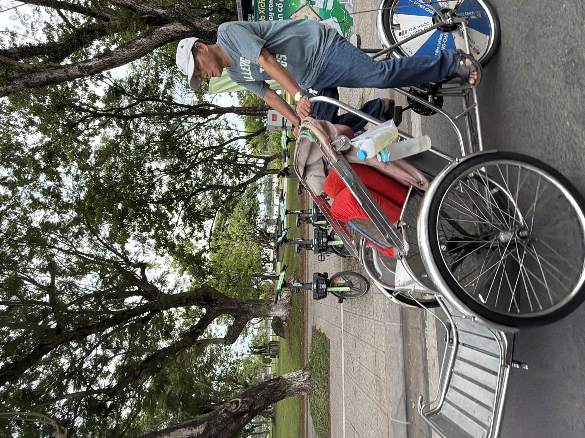A man is riding a cycle rickshaw on a paved road in Thuận Hóa Ward, Vietnam. He is wearing a light-colored baseball cap, a grey t-shirt with text, and blue jeans. The rickshaw has a passenger compartment with red and tan padding, and two water bottles are attached to the frame. In the background, there are several green electric scooters parked on a sidewalk next to a grassy area with trees and a statue. The overall scene appears to be a public park or a recreational area within the city.