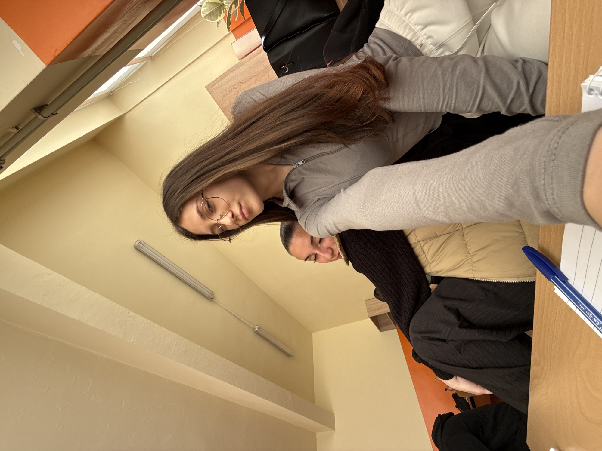 Two women are in a classroom. One woman is wearing glasses, a grey top, and has long brown hair. The other woman has dark hair, wears a black sweater, and a tan vest. There is a desk to the right with a blue pen and paper on it. The room is a yellow-orange color. There are fluorescent lights in the ceiling. The location is likely Sofia, Bulgaria.