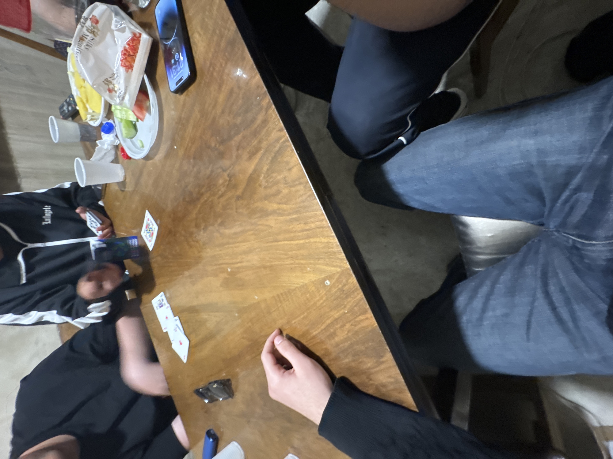 The image depicts a casual indoor gathering around a glossy wooden table in Yerevan, Armenia, during the evening. At least four individuals are present, with only parts of their bodies visible.
On the left side of the table, one person is wearing a black hoodie with white stripes and text that appears to be "L'Anglele" or "L'Angelo," holding a hand of playing cards. Below them, another person in a black long-sleeved shirt has a hand resting on the table near more playing cards. The right side of the table shows the legs of two other individuals: one in dark pants and dark shoes, and another in blue jeans.
The activity appears to be a card game or a social get-together. The table is laden with various items, including several playing cards, white plastic cups (some empty), and snacks. A white plastic bag, partially showing the text "Silk," contains what look like chips or puffed snacks. There is also a small plate with a salad of lettuce, tomato, and cucumber, alongside some yellow food items. A mobile phone, likely an iPhone, is placed face-up, displaying the time "21:31." Other small objects include a blue bottle cap, a small black rectangular item (possibly a lighter), and a blue pen or marker. The dim lighting suggests a relaxed, informal evening atmosphere.
