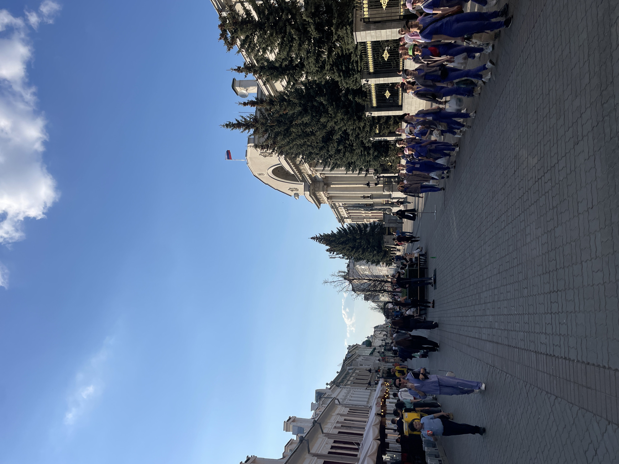 The image depicts a street scene in Kazan, Russia. On the left is a light-colored building with columns and a Russian flag on top. Next to the building is a large evergreen tree. A line of people, mostly in blue uniforms, are standing along the right edge of the street. There is a row of buildings in the lower part of the image with storefronts and people walking along the street. The sky is mostly blue with some white clouds. The street is paved with grey paving stones.