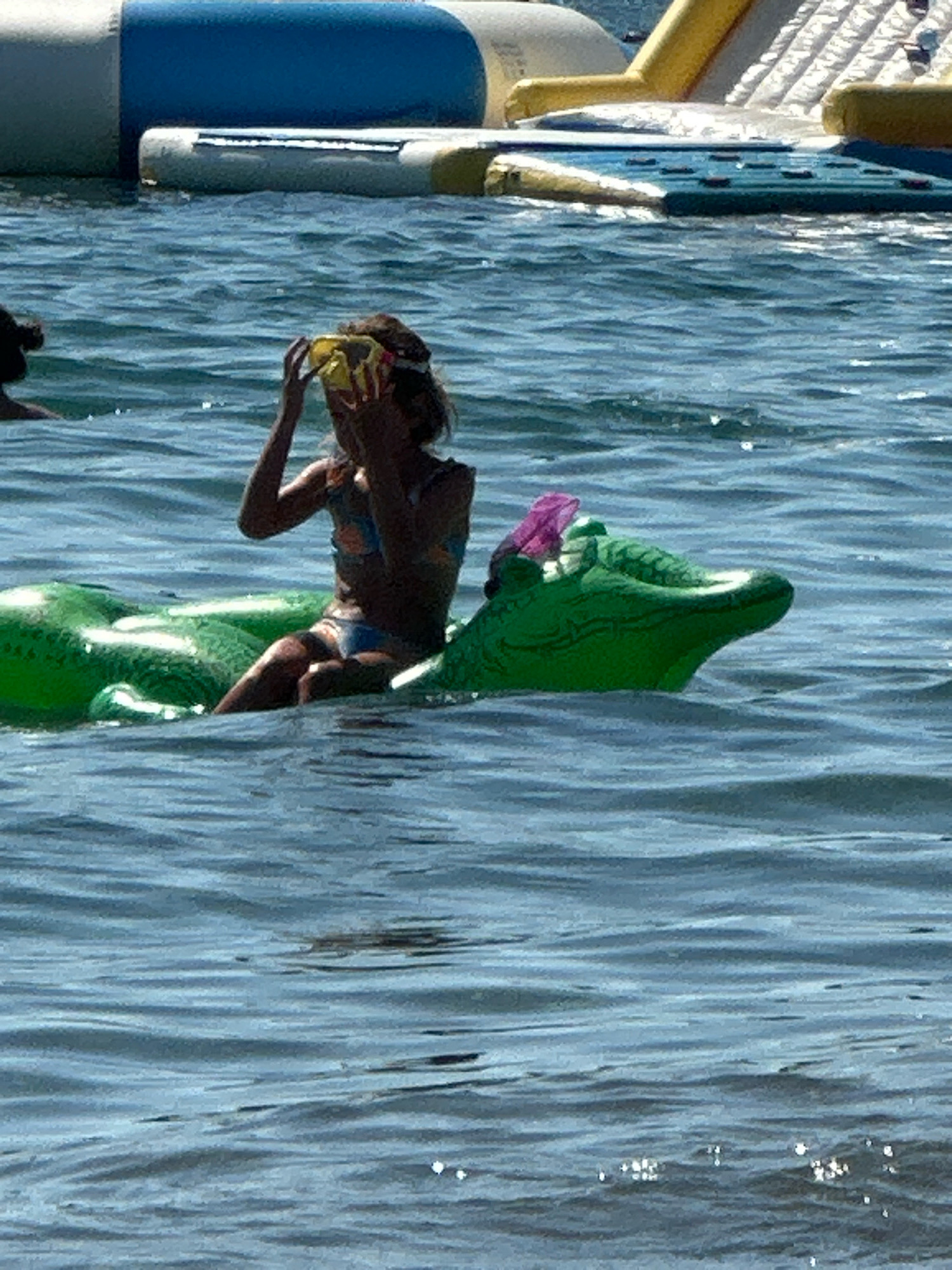 In the sparkling waters of Boreti, Montenegro, a young girl is captured mid-activity. She is seated on a bright green inflatable alligator, her snorkel mask pushed up to her forehead. The girl, wearing a colorful bikini, is focused on something off-camera, her hands near the mask. The water is a deep blue, rippled by a gentle breeze, suggesting a pleasant day. In the background, a large inflatable water park structure, featuring a blue tube and yellow slide, hints at a recreational area. The overall scene conveys a sense of summer fun and outdoor enjoyment, with the sun likely high in the sky, casting bright reflections on the water. No visible text is present in the image.