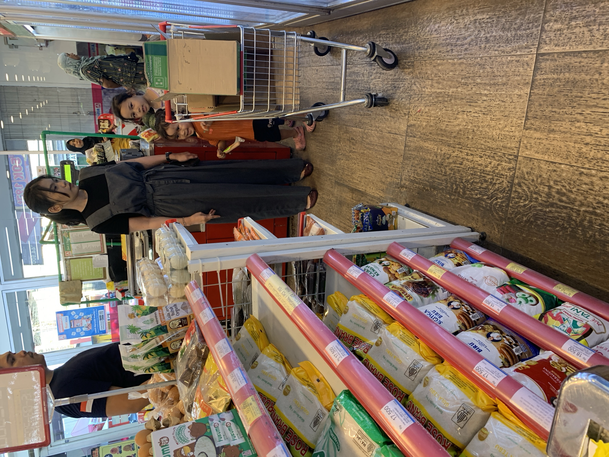 In a bright grocery store aisle, a woman in a gray apron stands near a shopping cart filled with items. Two young girls are in the cart with her, one looking at the camera and the other holding onto a shopping cart handle. In the background, a man in a black t-shirt and a woman wearing a hijab are visible, along with shelves stocked with various products like bags of sugar, rice, and other groceries. The flooring is made of brown tiles, and the overall setting suggests a commercial establishment in Makassar, Indonesia.