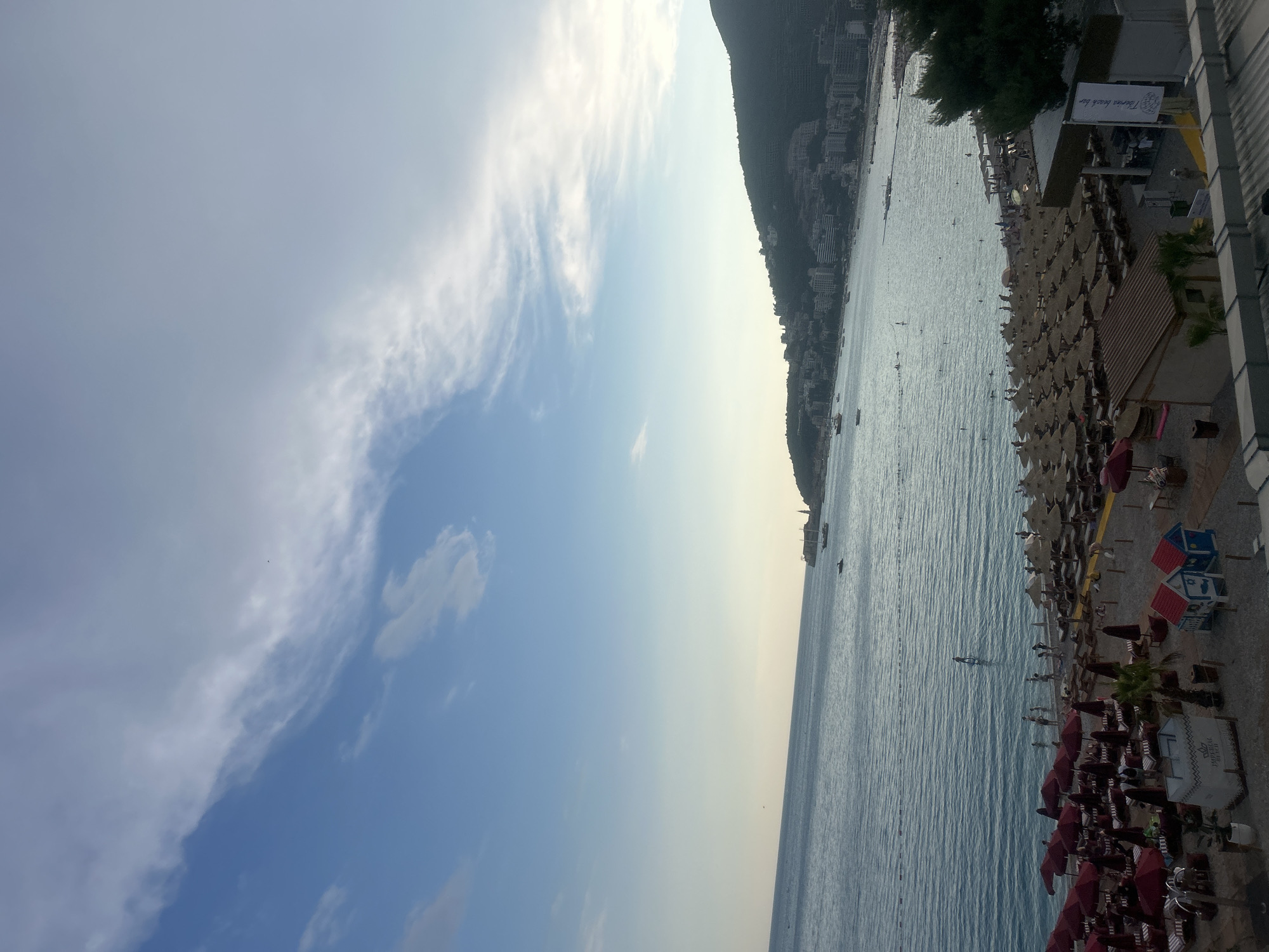 This is a vertical, eye-level shot of a bustling beach scene in Budva, Montenegro. The image captures a vibrant stretch of coastline, with the turquoise Adriatic Sea on the left and a densely populated beach on the right.
The sea is calm, with small waves gently lapping at the shore. Scattered across the water are several boats, some appearing to be moored, while others might be in use. A person is visible swimming in the water, adding to the sense of activity.
The beach is packed with beach chairs and umbrellas, indicating a popular day. Rows of straw umbrellas offer shade to sunbathers, and some small beach huts are present. People are scattered amongst the chairs and walking along the sand.
Dominating the background on the right is a steep, tree-covered mountain, with buildings densely packed along its slopes, characteristic of Budva's coastal landscape. The sky is a pale blue with wispy white clouds, suggesting it is daytime, likely late afternoon given the warm, diffused light.
Visible text includes a sign on the beach that reads "Turism Beach Bar". The overall atmosphere is one of a lively, sunny day at the beach.