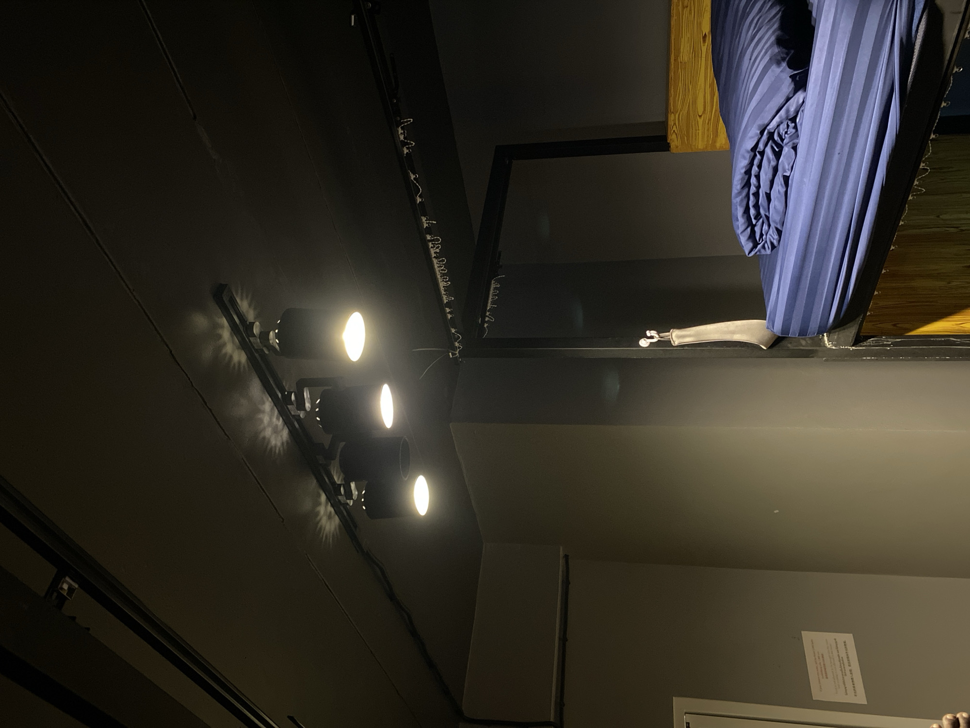 The image captures an indoor scene, likely a modern or compact living space, viewed from a low angle looking upwards. The primary lighting comes from three bright, circular spotlights mounted on a track on a dark ceiling, casting starburst patterns of light.
Above these lights, an elevated structure or loft bed is visible. A tightly rolled or folded blue, striped fabric, resembling a mattress or duvet, hangs down from this upper platform. To the left of the blue fabric, a wooden beam provides support for the elevated area. Below the blue fabric and resting on the edge of the elevated platform is a distinctive object: a pale, jointed prosthetic-like leg or doll's leg, featuring small, wheel-like attachments at its end.
The walls and ceiling are painted in dark, muted tones, possibly dark grey or black, creating a somewhat enclosed atmosphere. Along an upper beam or track on the left, a string of small decorative lights or fairy lights is visible. On a lighter-toned wall in the lower right quadrant of the image, a white rectangular object, possibly an air conditioning unit or vent, is present. Below this, a white piece of paper with densely printed text is taped to the wall. While largely illegible, the text appears to be a notice or instructions, possibly in multiple languages including English (with words like "Lounge" or "Laundry" faintly visible) and an Asian script, consistent with the given location context of Pattaya City, Thailand. No people are visible, and no specific activity is taking place in this static scene. The artificial lighting suggests it could be night or an interior space with limited natural light.